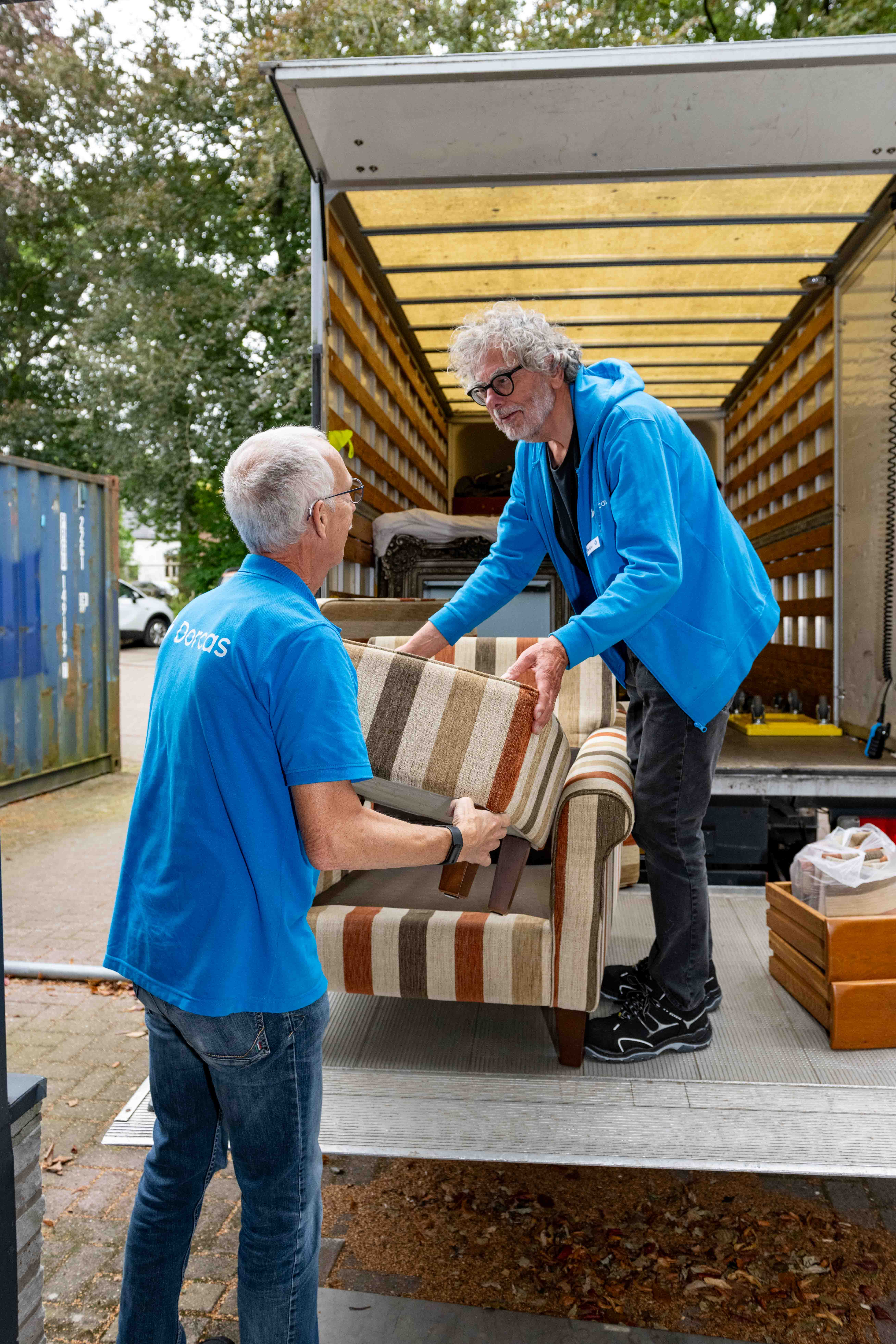 Twee vrijwilligers van Dorcas helpen een bank uit een verhuiswagen te halen en zetten deze voorzichtig op de grond.