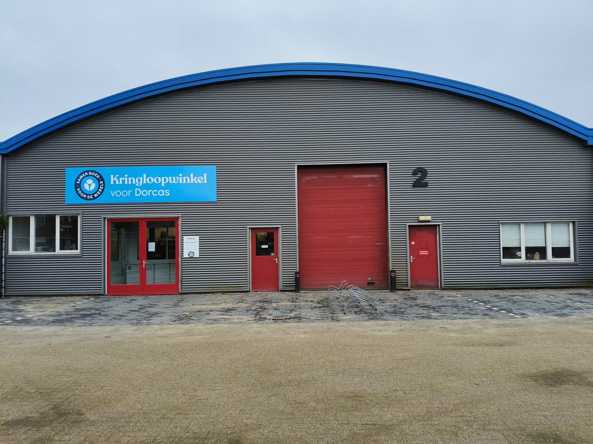 Kringloopwinkel voor Dorcas in Aalten, gevestigd in een grote, grijze bedrijfsloods met blauwe en rode accenten, biedt tweedehands goederen en donaties.