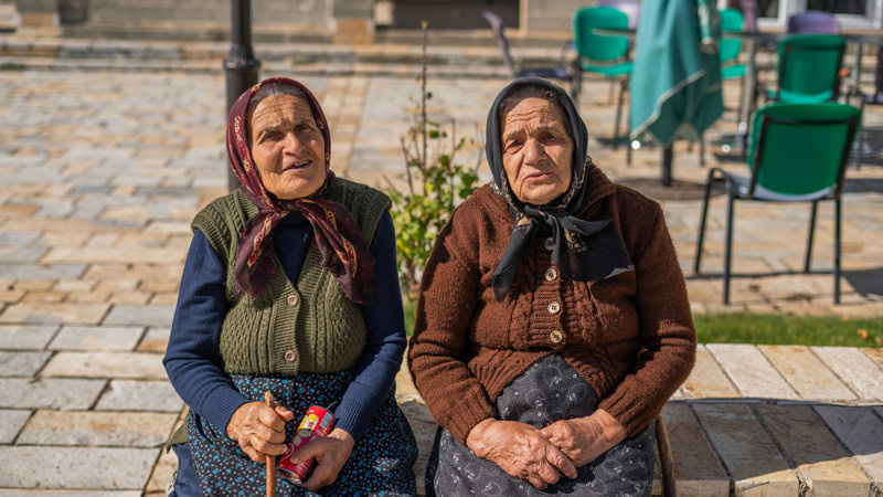 Een foto van twee oudere vrouwen in Albanië die samen buiten zitten, met een rustige en vriendelijke uitstraling, onder een heldere hemel.