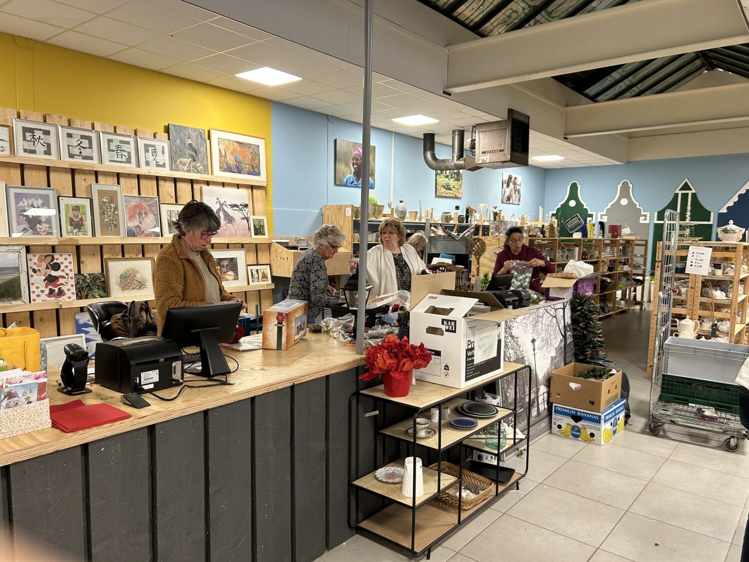 Mensen shoppers in een tweedehands winkel met gekleurde muren, kunstwerken aan de muur en diverse artikelen op de kassa en schappen.