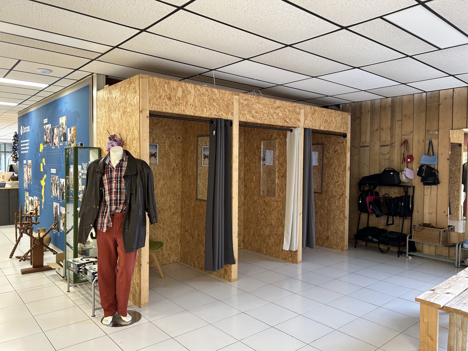 Dorcashol wordt zichtbaar, met paspop gekleed in jas en broek en koopjeshoek met gordijnen voor privé paskamers.