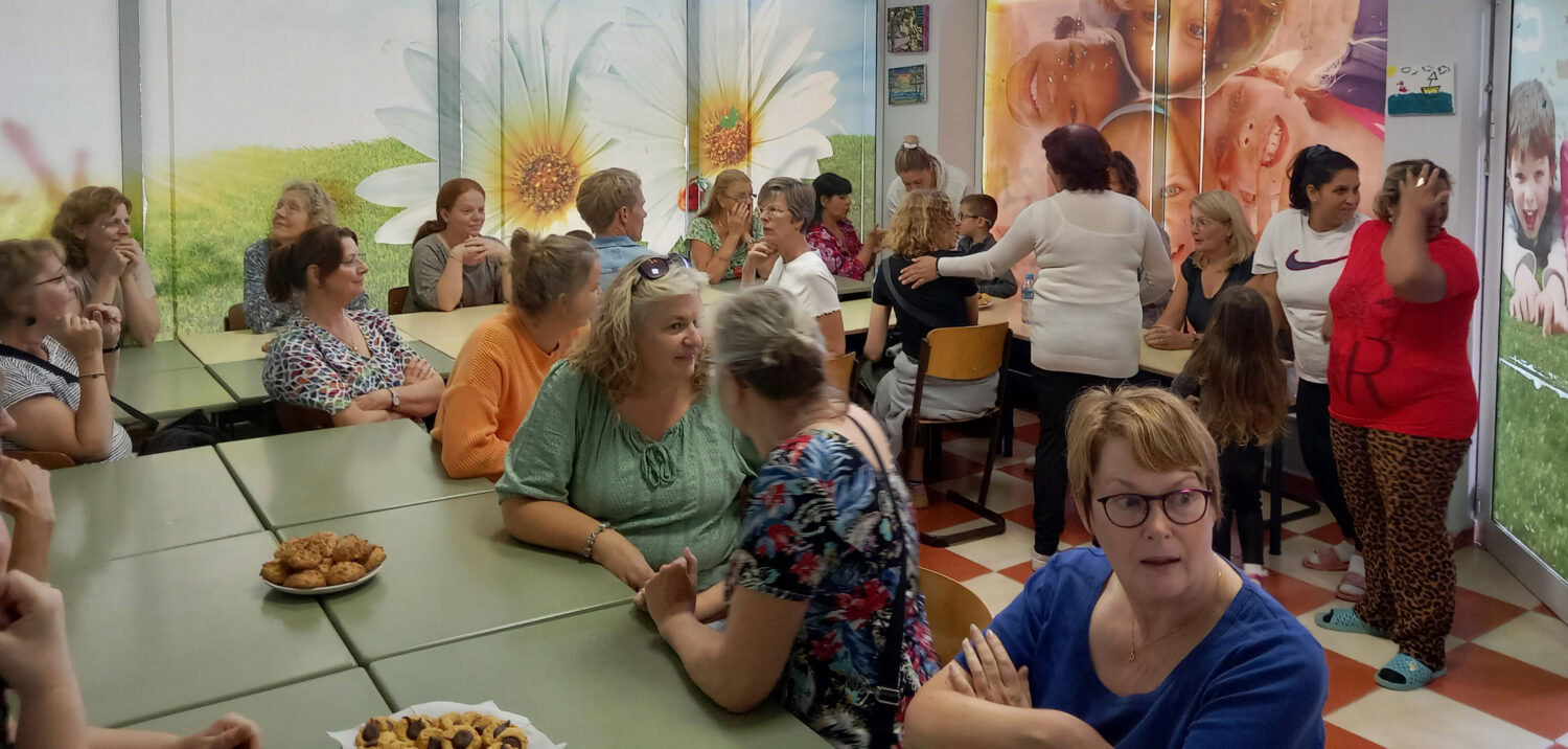 Gezellig bijeenkomst met mensen die in gesprek zijn, in een kleurrijke ruimte met muurdecoraties van bloemen en lachende kinderen, georganiseerd door Dorcas.
