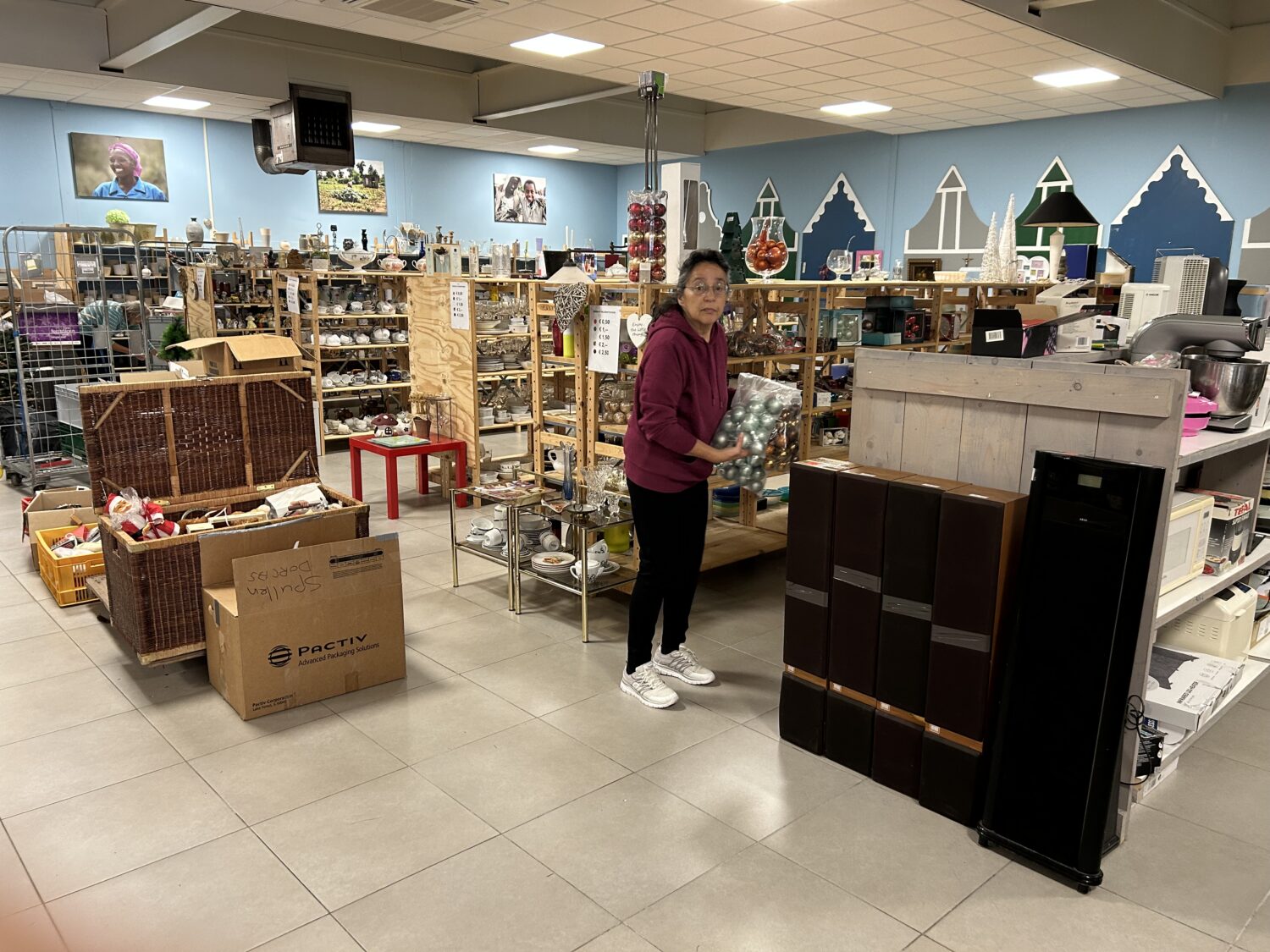 Vrouw die kerstballen vasthoudt in een kringloopwinkel met verschillende tweedehands spullen en decoraties op houten planken.