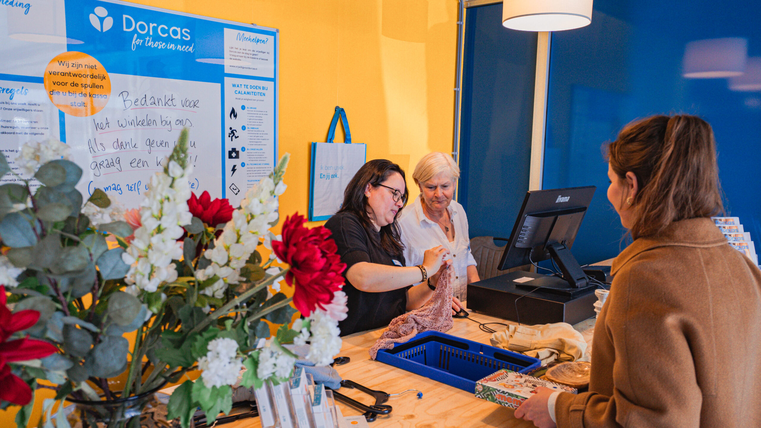 Drie vrouwen aan de balie in de Dorcas winkel, betrokken gesprek over vrijwilligerswerk en inkoop, met bloemen op de voorgrond en informatiemateriaal op de achtergrond.