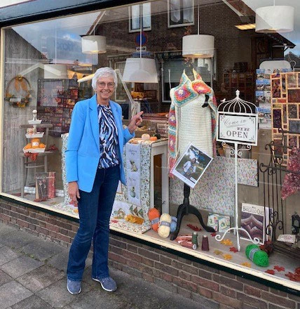 Gezellige vrouw voor winkelraam met handgemaakte handwerken, breiwerk en decoraties, winkel van Dorcas met open sign in het raam.
