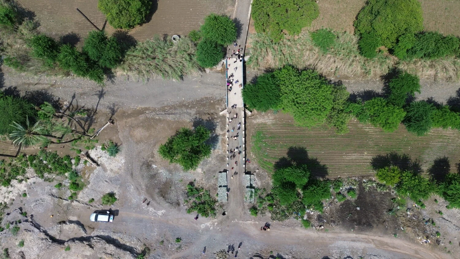 Gezicht vanaf boven op een smalle brug met mensen die erover lopen, omgeven door groene bomen en akkers, met een auto en een ruwe pad onder de brug.
