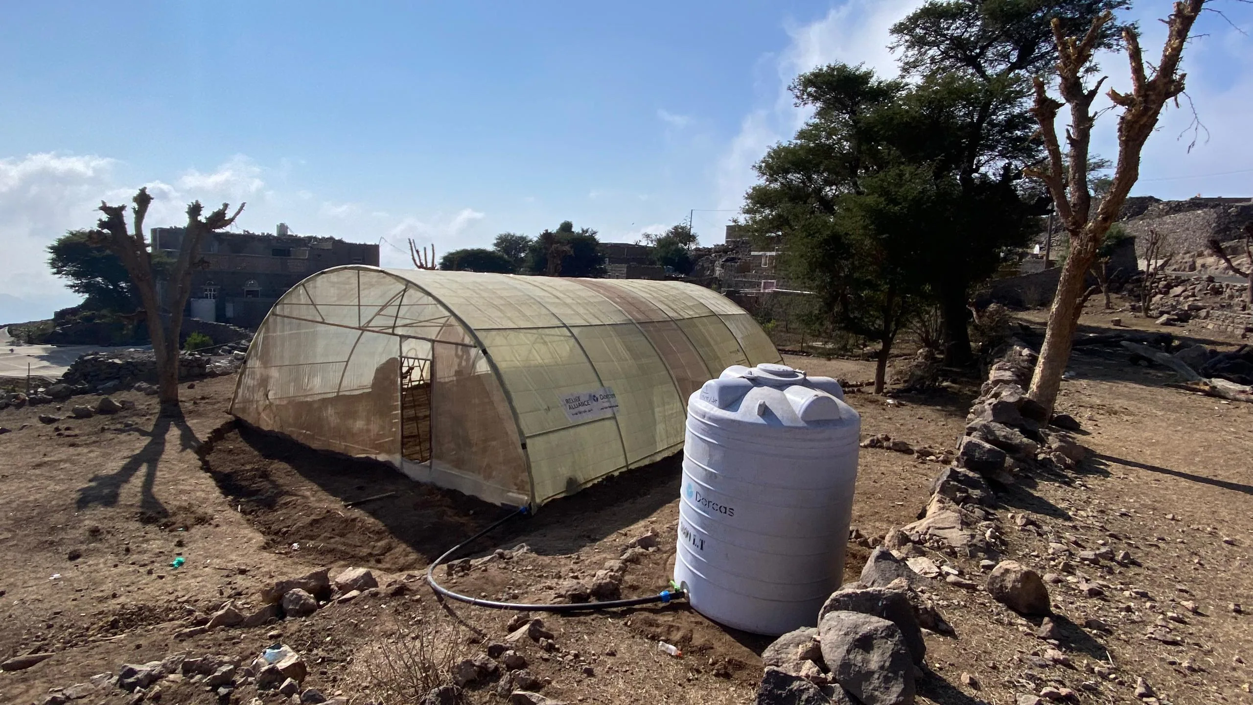 Kleine kas met uitstalraam, geïnstalleerd op een drooge, steenrijke grond met stenen en bomen op de achtergrond, voor duurzame landbouw of tuinprojecten in Afrika.