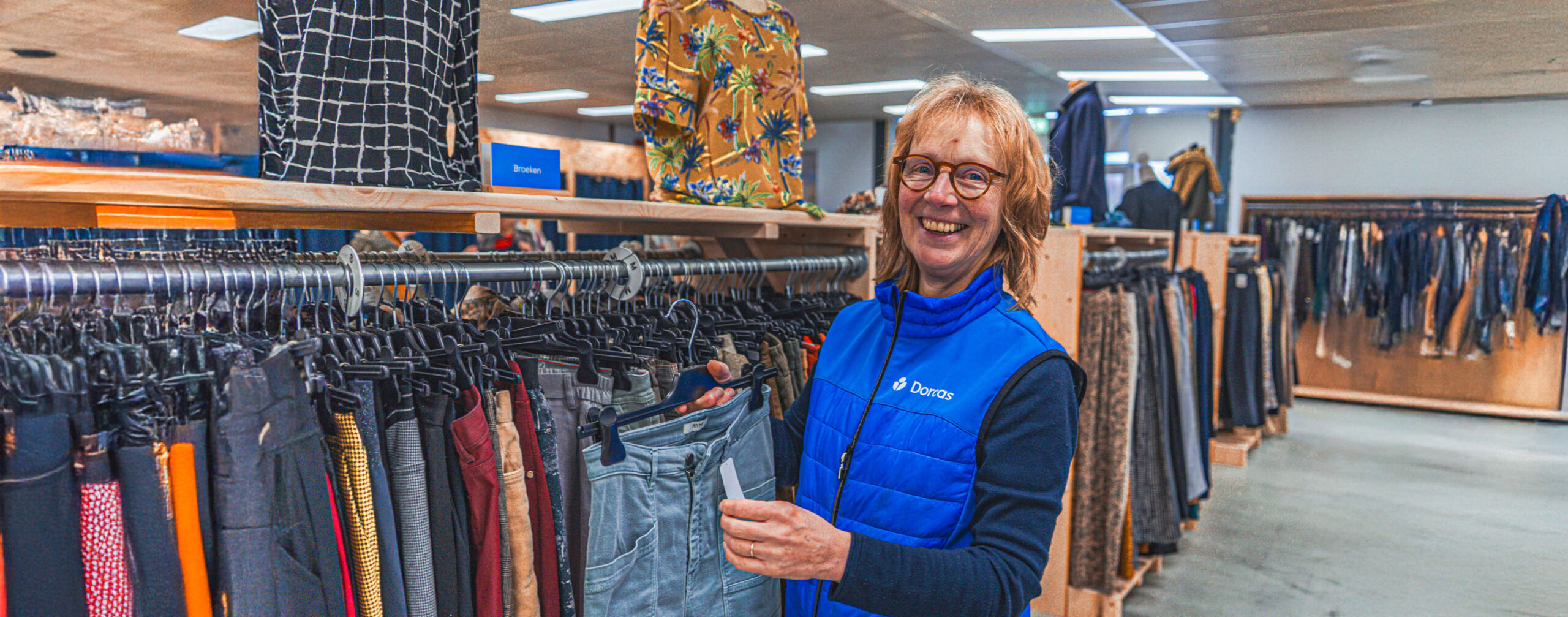 Dame met winkelmedewerker in Doris winkel, die kleding uitkiest op een kledingrek met diverse outfits en accessoires in een heldere winkelruimte.