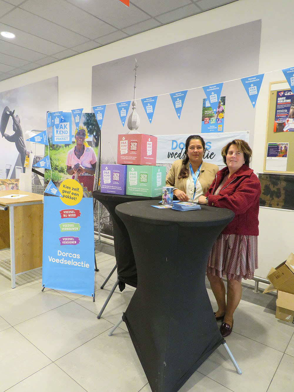 Kraam op een Dorcas voedselactie met twee vrouwen en promotiekleding, posters en flyers over voedselhulp en verbeelding van Dorcas projecten, binnen in een winkelcentrum of evenementruimte.