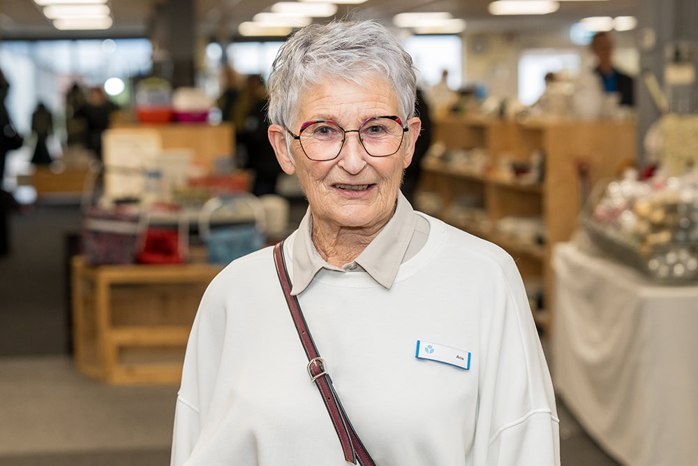 Oudere vrouw met brillen en naamkaartje in winkelvolledig met houten rekken en klanten op de achtergrond.