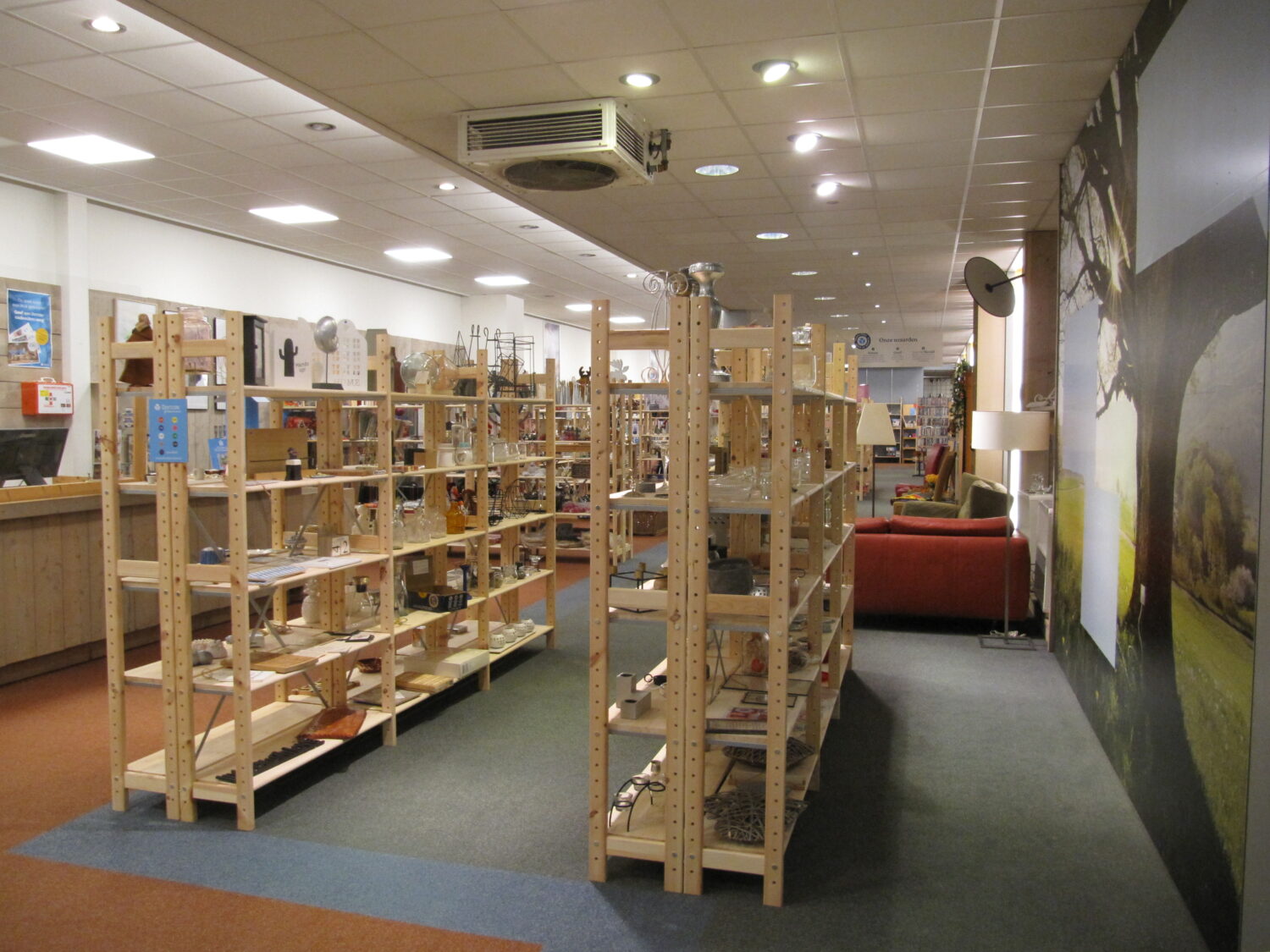 Dorrecas' winkel met houten rekken vol tweedehands spullen, gezellige zithoek met lampen en grote muurprint, promotie voor duurzaam winkelbezoek en hergebruik.