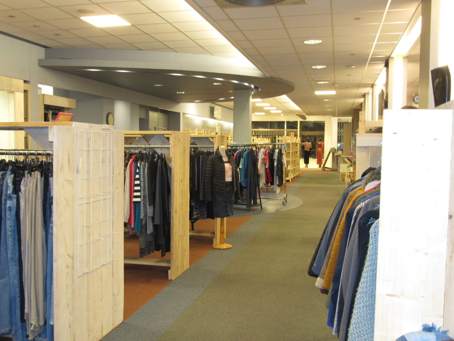 Duurzaam winkelbeeld met kledingrekken vol tweedehands mode, houten winkelinrichting en ruime winkelgang in het interieur van een sociaal mode-atelier van Dorcas.