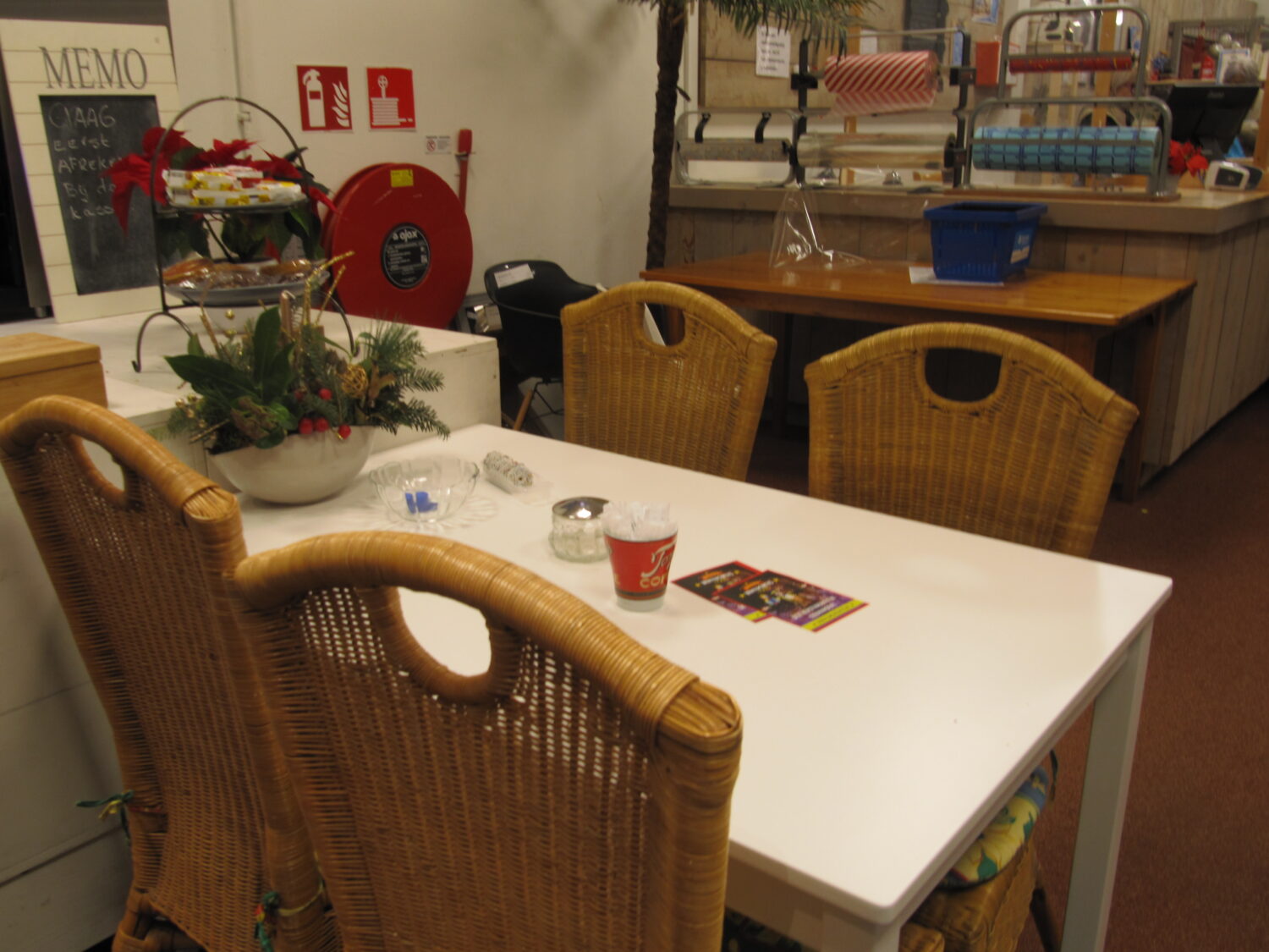 Kerstdecoratie op een wit tafel, naast rieten stoelen, in een gezellig winkelinterieur met winkelrekken en verpakkingsmateriaal op de achtergrond.