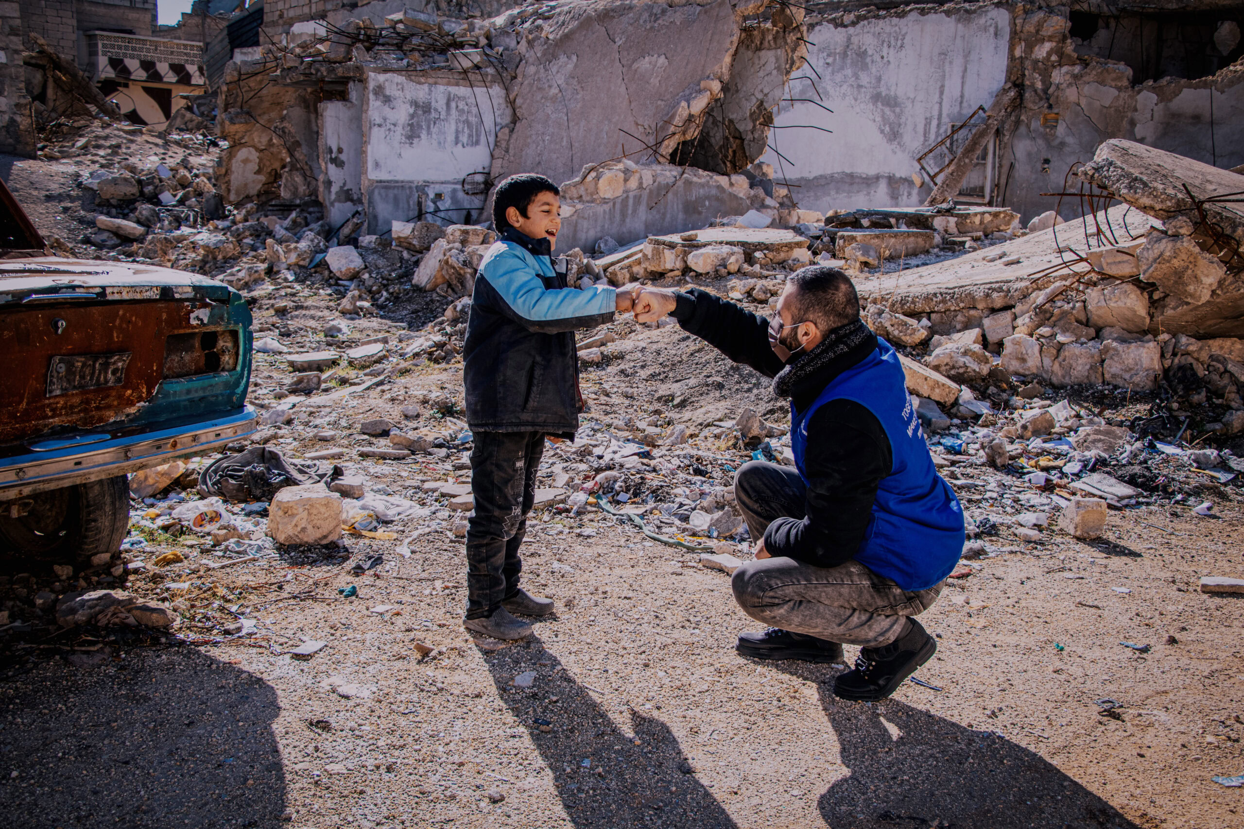 Hulpverlener geeft een kind een hand in een verwoeste omgeving na een natuurramp of conflict.