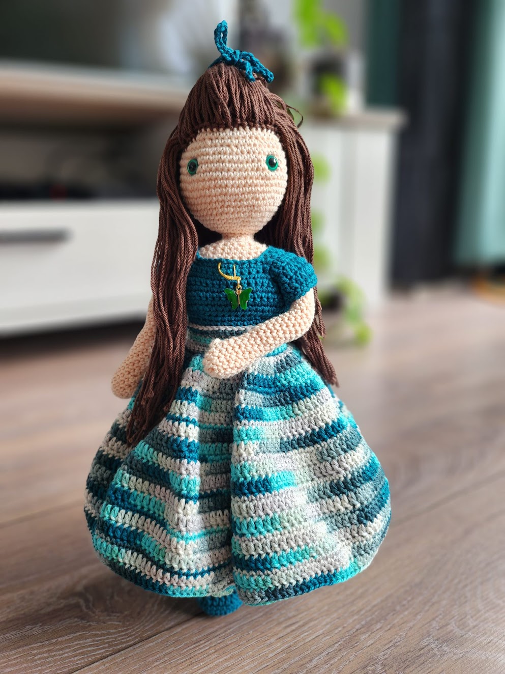 Kleurrijk gehaakte pop met lange bruin haar, blauwe top en jurk, detail van een groene vlinderschatketting, zittend op een houten vloer in een gezellige woonomgeving.
