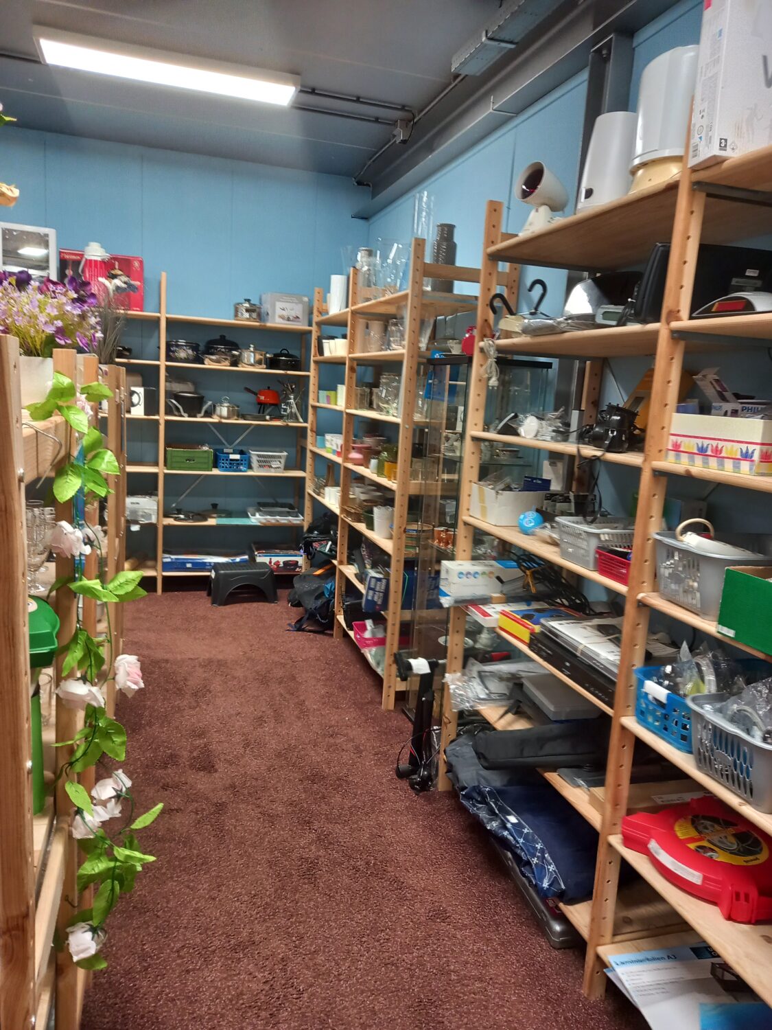 Duurzame tweedehandswinkel met uitgebreide collectie huishoudelijke artikelen en accessoires op houten schappen, in een gezellige en geordende sfeer voor iedereen die budgetvriendelijk wil shoppen.