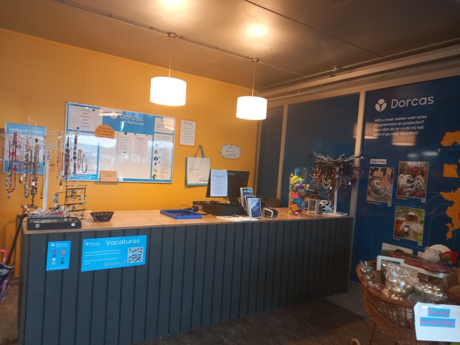 Dorcas inloopbalie met display en cadeaus, kleurrijke stenen en informatie over projecten, in een gezellige kantoorruimte met gele muur en blauwe wand.