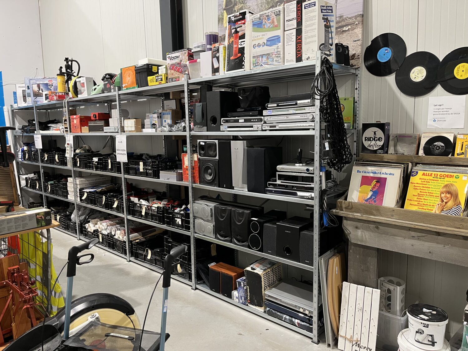 Veel elektronica en audioapparatuur op metalen rekken bij Dorcas tweedehandswinkel.