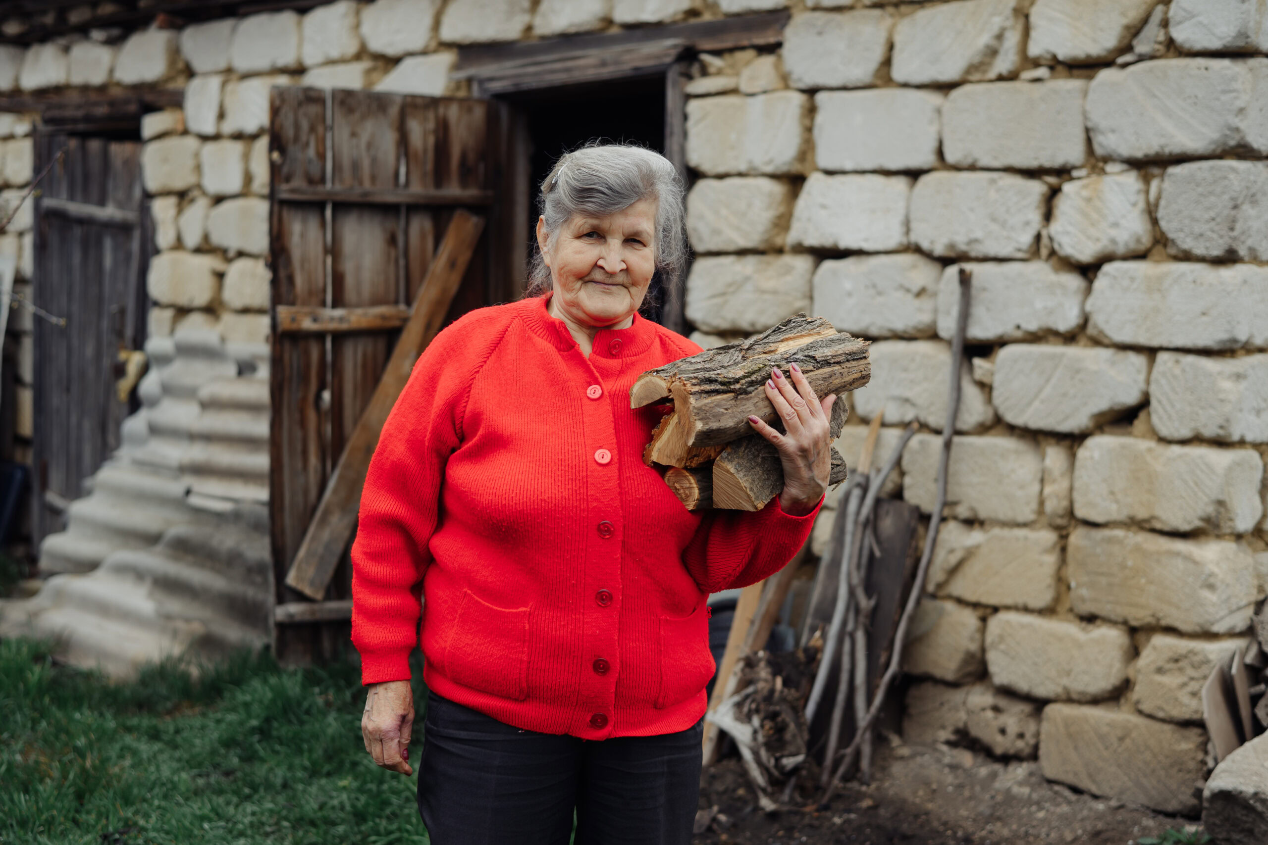 Kleine oude vrouw met grijze haar in een rode trui, die hout draagt buiten een erf met stenen muur en schuur.