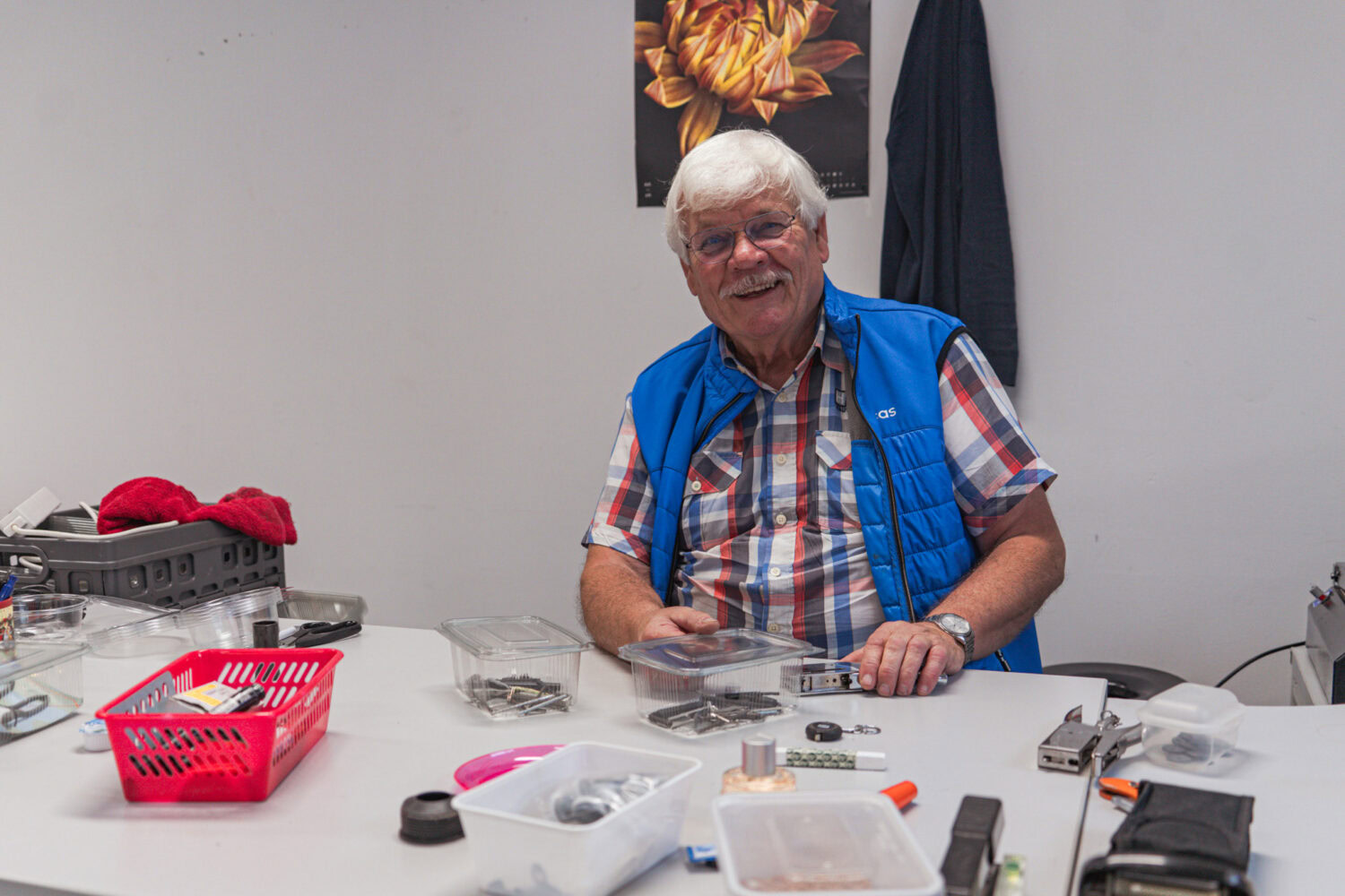 Een glimlachende oudere man met witte haar en bril, zittend aan een tafel vol gereedschap en accessoires, in een kantoor of werkruimte.