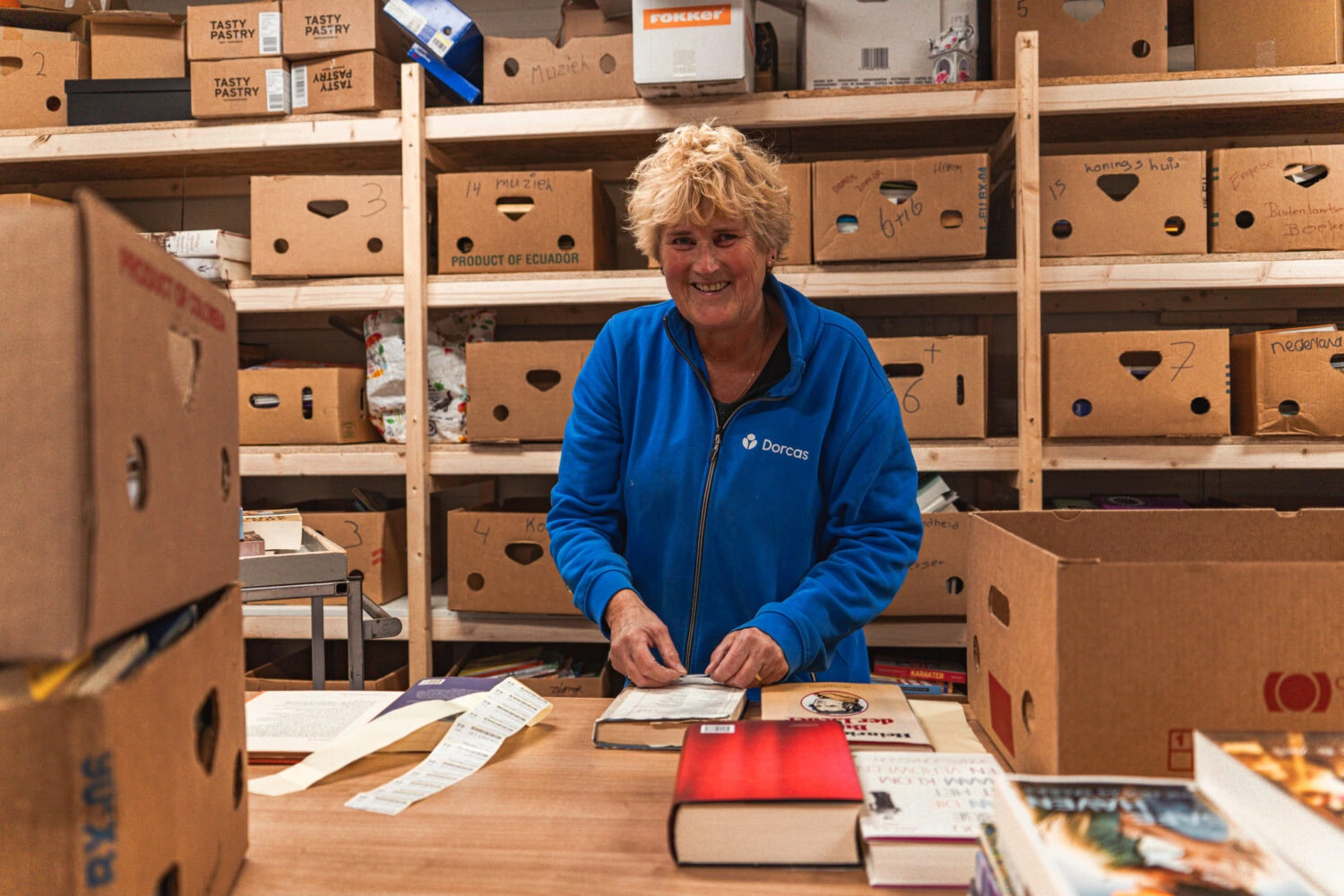 Dame in magazijn van Dorcas met dozen, boeken en vrijwilligers, organisatie voor armoedebestrijding en kledinginzameling in Nederland.