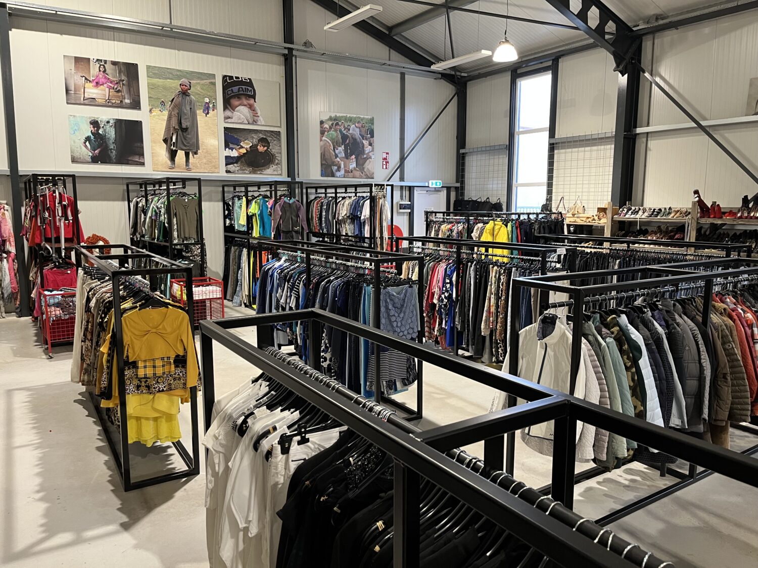 Dame winkelinrichting met kledingrekken vol kleding, accessoires en schoenen, in een industriële winkelruimte met hoge plafonds en grote ramen.