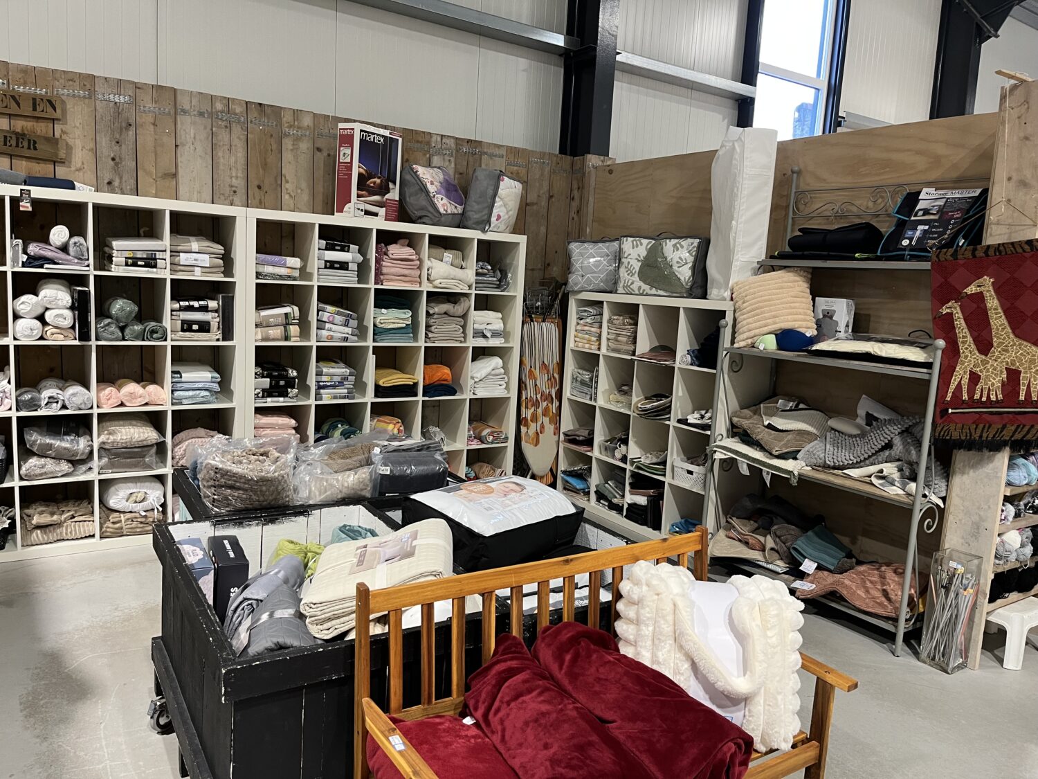 Dichte winkelvloer met wit houten rekken gevuld met textiel, dekens, kussens en huishoudartikelen, vooral in pastel- en aardetinten, met natuurlijke hout- en metalen details.