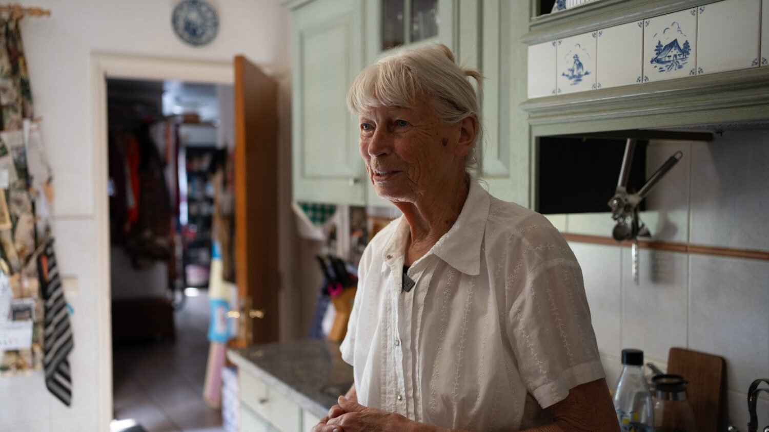 Kleine oude vrouw in de keuken, veel natuurlijk licht, groene vintage keukenkastjes, gezellig en huiselijk interieur, dagelijks leven.