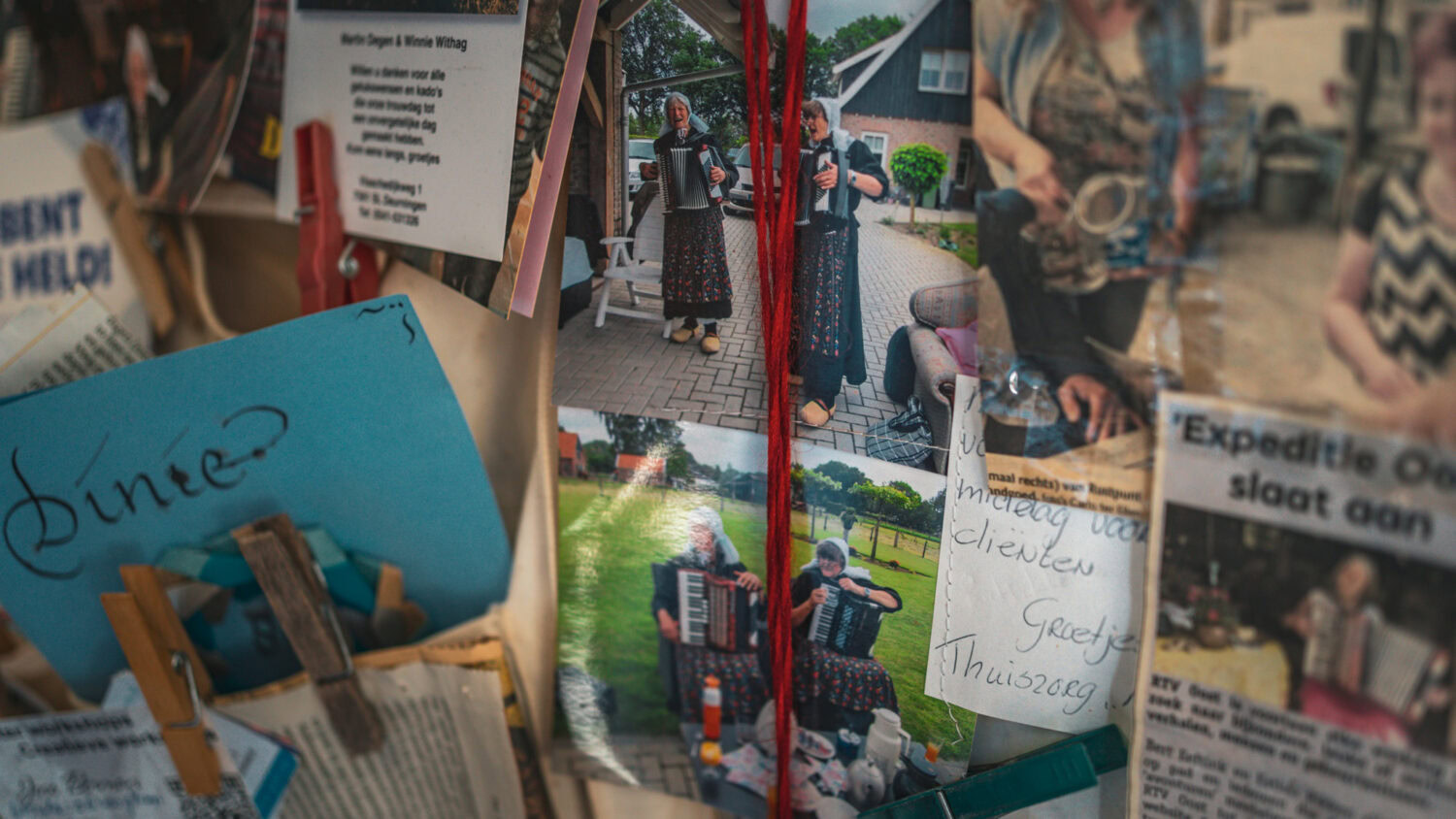 Gezicht van twee vrouwen die accordeon spelen op een buitenmarkt, omringd door foto's en notities, met een huis en bomen op de achtergrond.