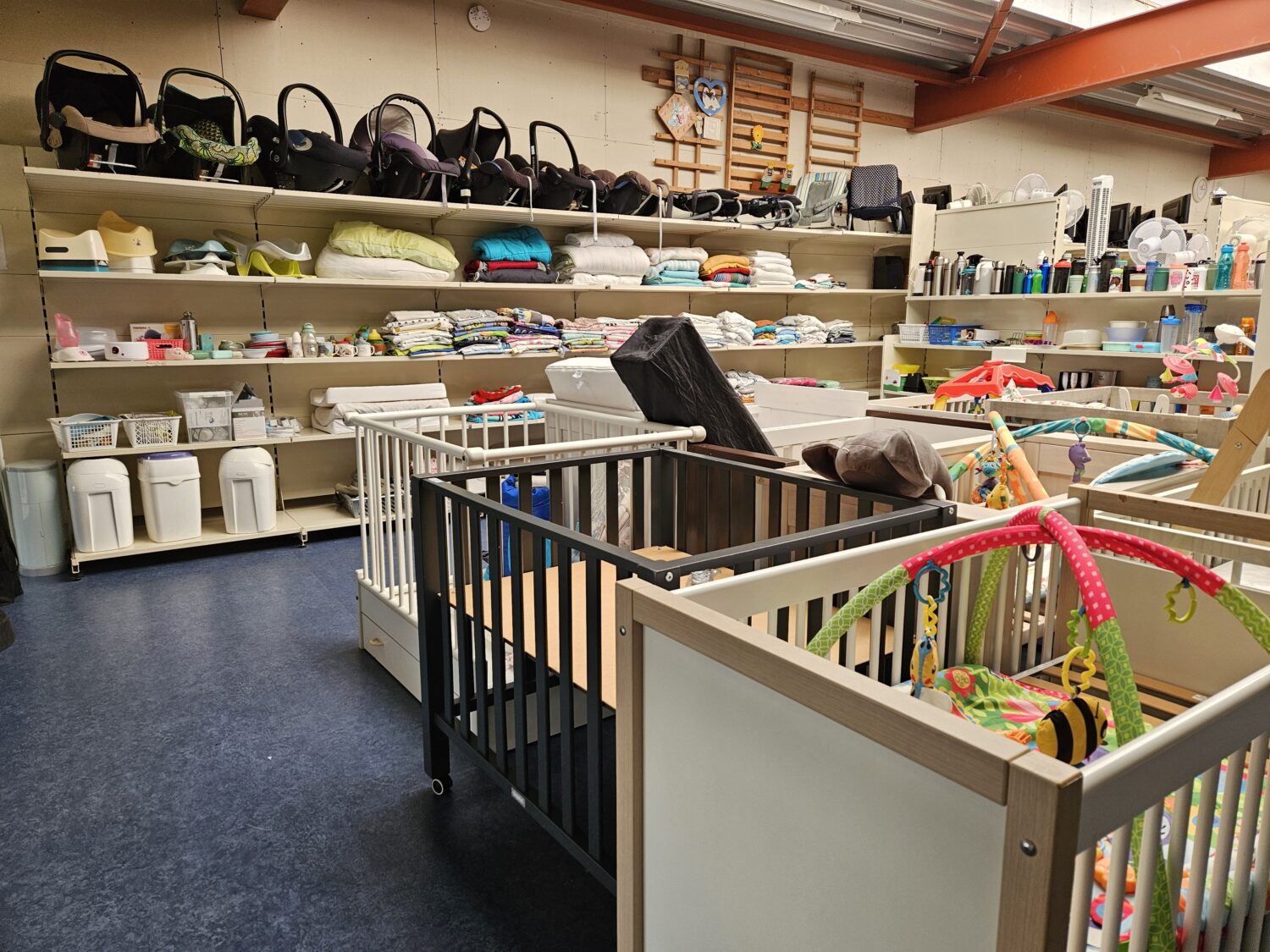 Een overzicht van diverse kinderwagens, babyspullen en accessoires in een tweedehandswinkel, netjes uitgestald op planken en in kinderbedjes, klaar voor verkoop.