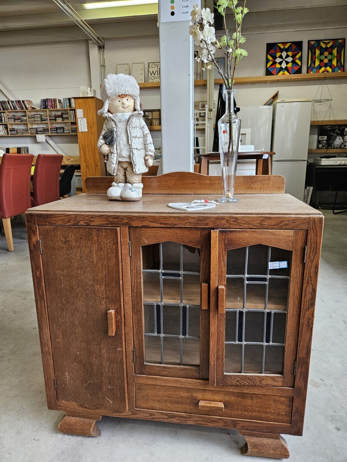 Een antieke houten kast met glazen deuren, versierd met een vaas met witte bloemen en een knuffelpop, geplaatst in een tweedehandswinkel of antiekzaak.