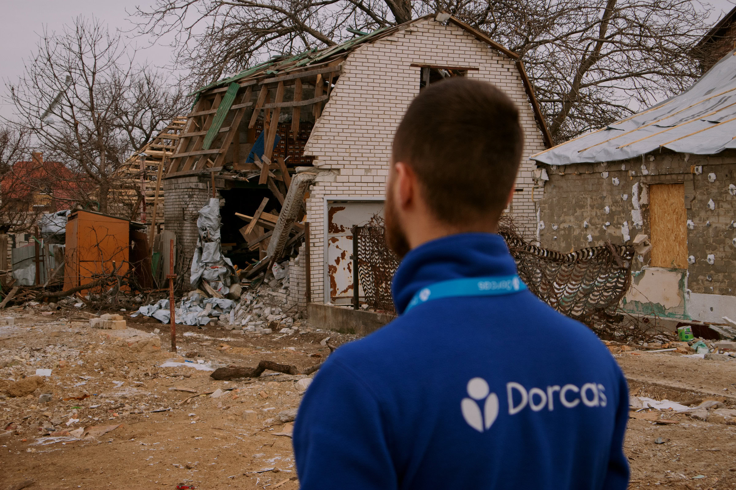 Een man met een blauwe Dorcas-jas kijkt naar een verwoest huis dat beschadigd is door de oorlog in Oekraïne, symboliserend de hulp en wederopbouw door Dorcas.