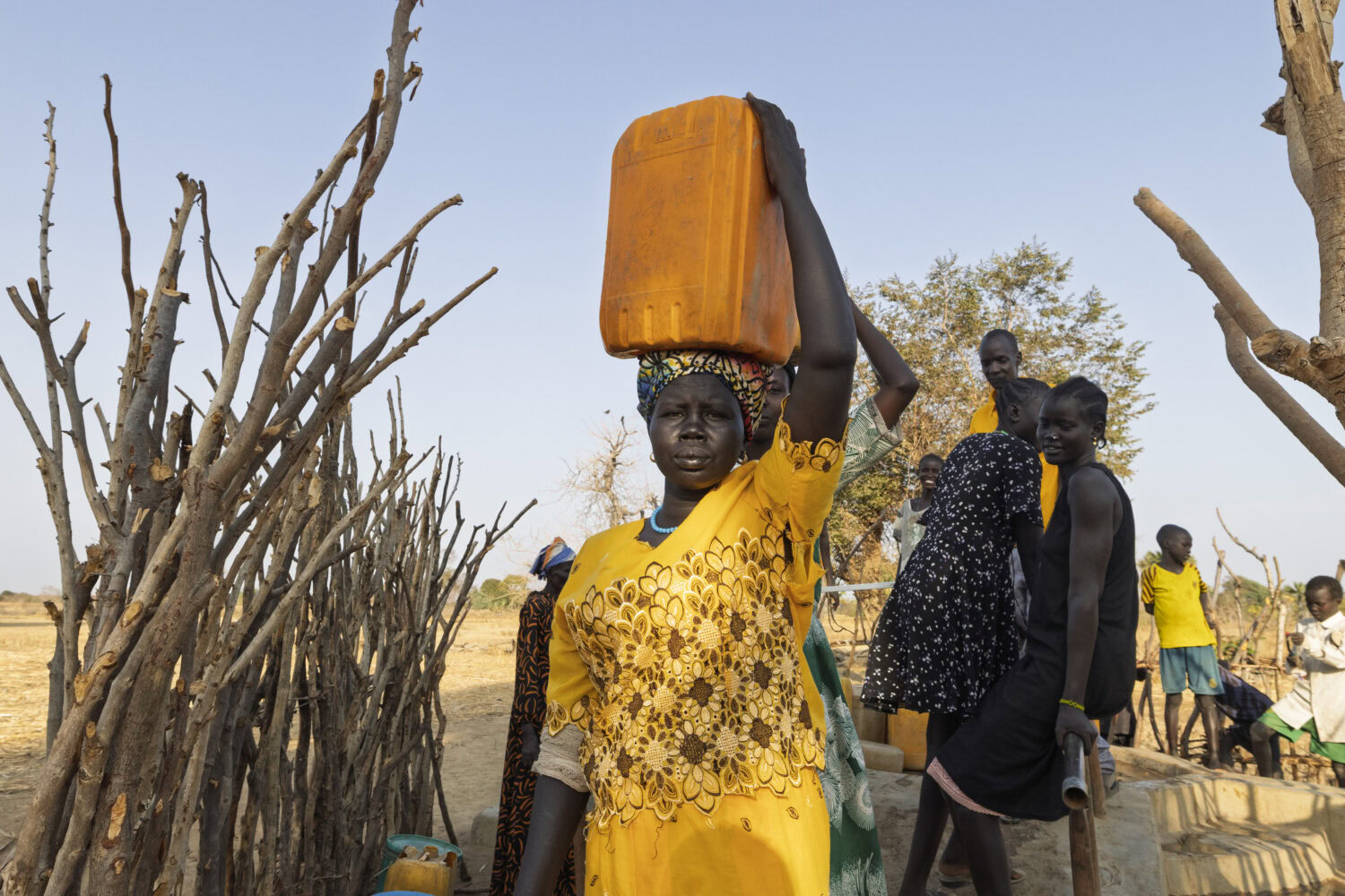 Vrouw in geel kleding draagt grote watercontainer op haar hoofd, omringd door andere mensen in een dorre, Afrikaanse omgeving.
