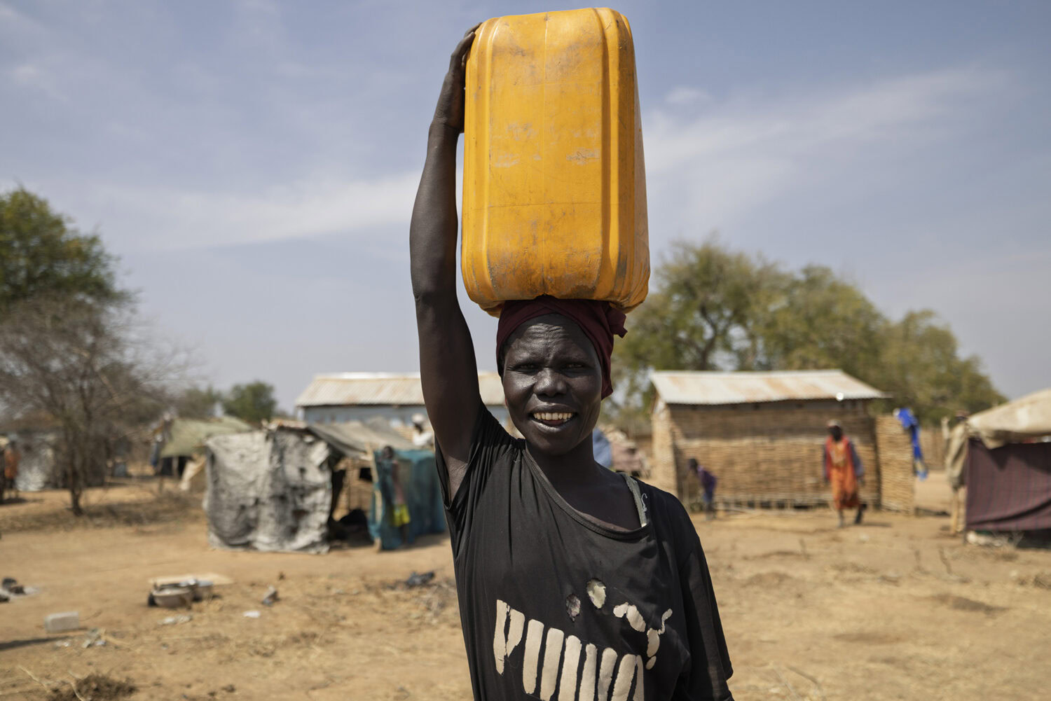 Een vrouw in een droog landschap met een grote gele container op haar hoofd, symboliseert waterkracht en hulpverlening in actie.