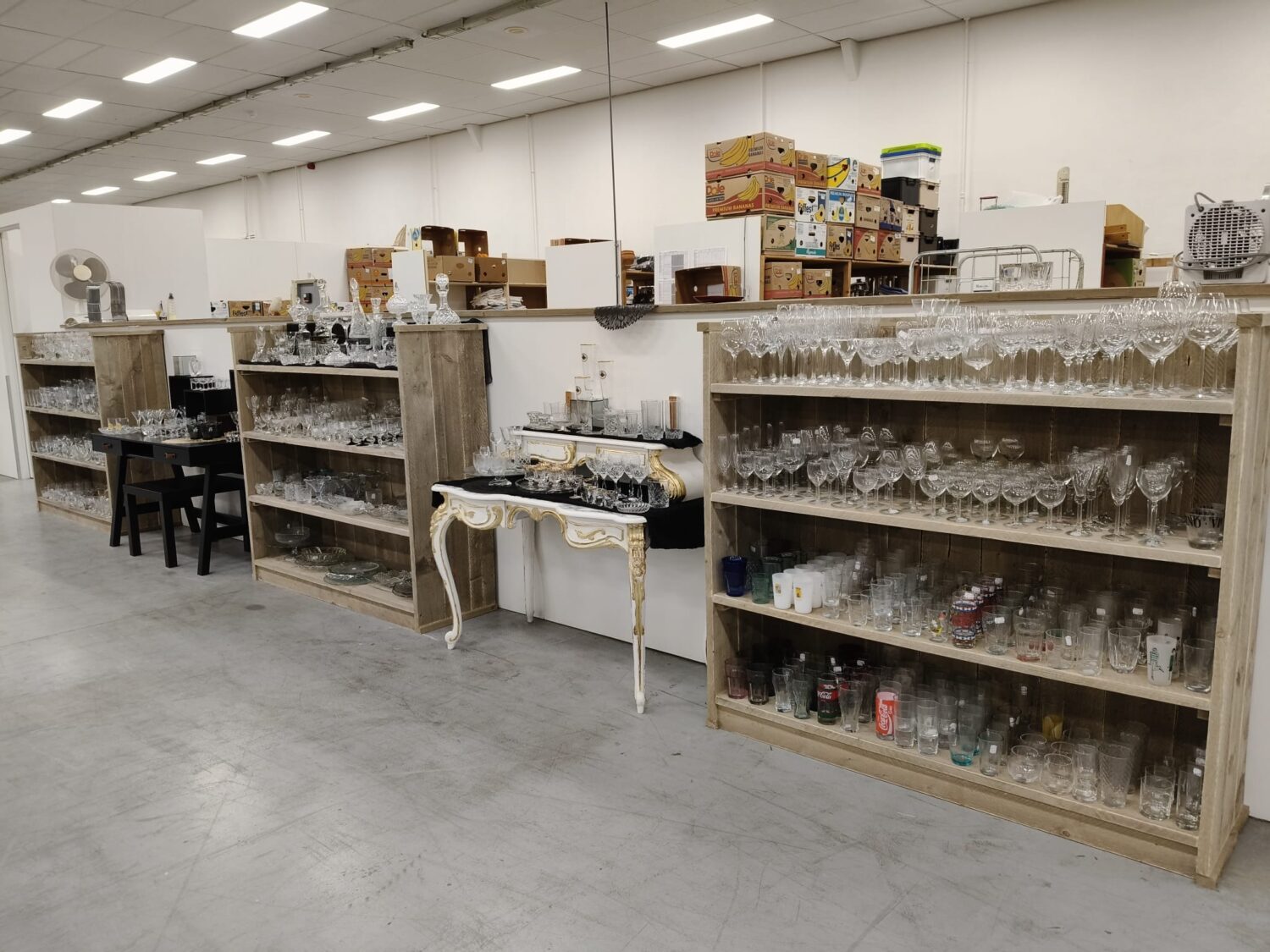 Een overzicht van glazen, servies en andere huishoudartikelen op houten planken in een winkel of opslagruimte, klaar voor verkoop of distributie.