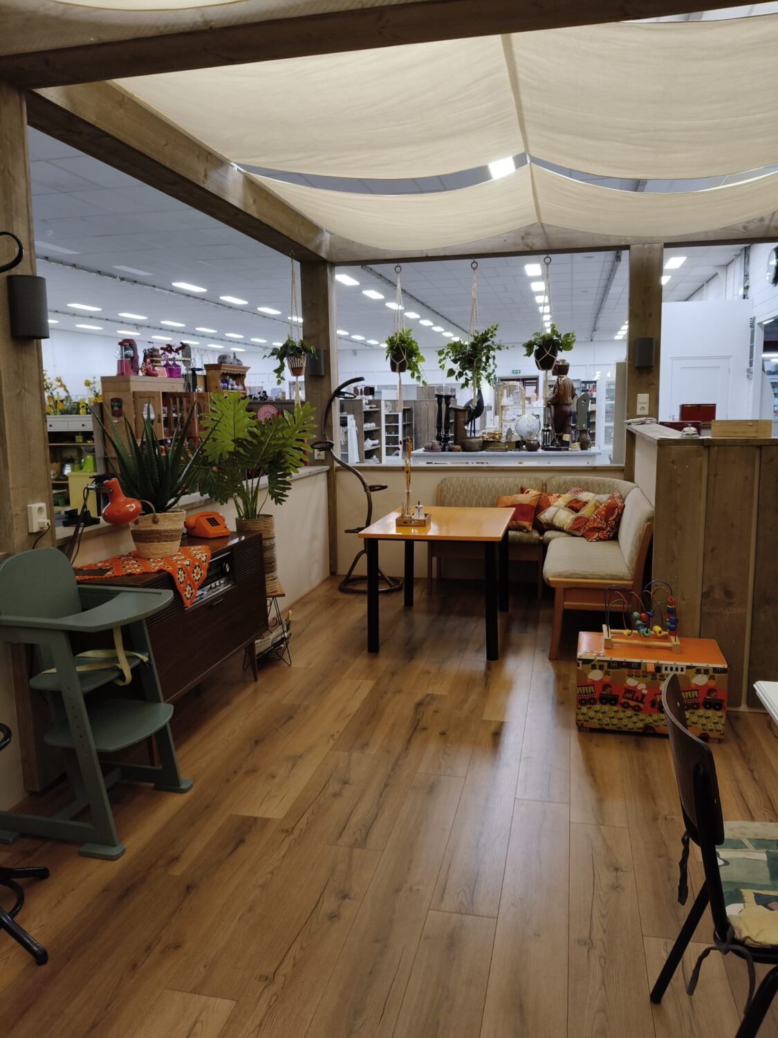 Gezellige zithoek met planten en comfortabele bank, omringd door winkelvoorraad en decoratie, binnen een sfeervolle winkelruimte met houten vloer en grote ramen.