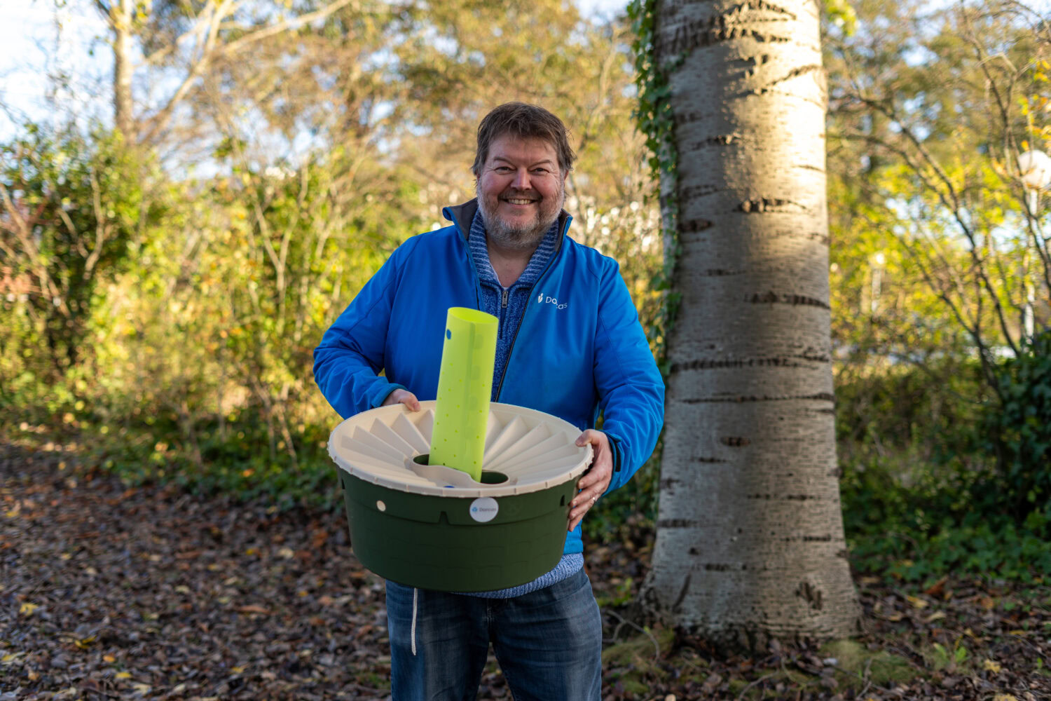 Man met een grote glimlach houdt een groene en witte container met een grote groene pen erin, buiten in een bosrijke omgeving met bomen en herfstkleuren.