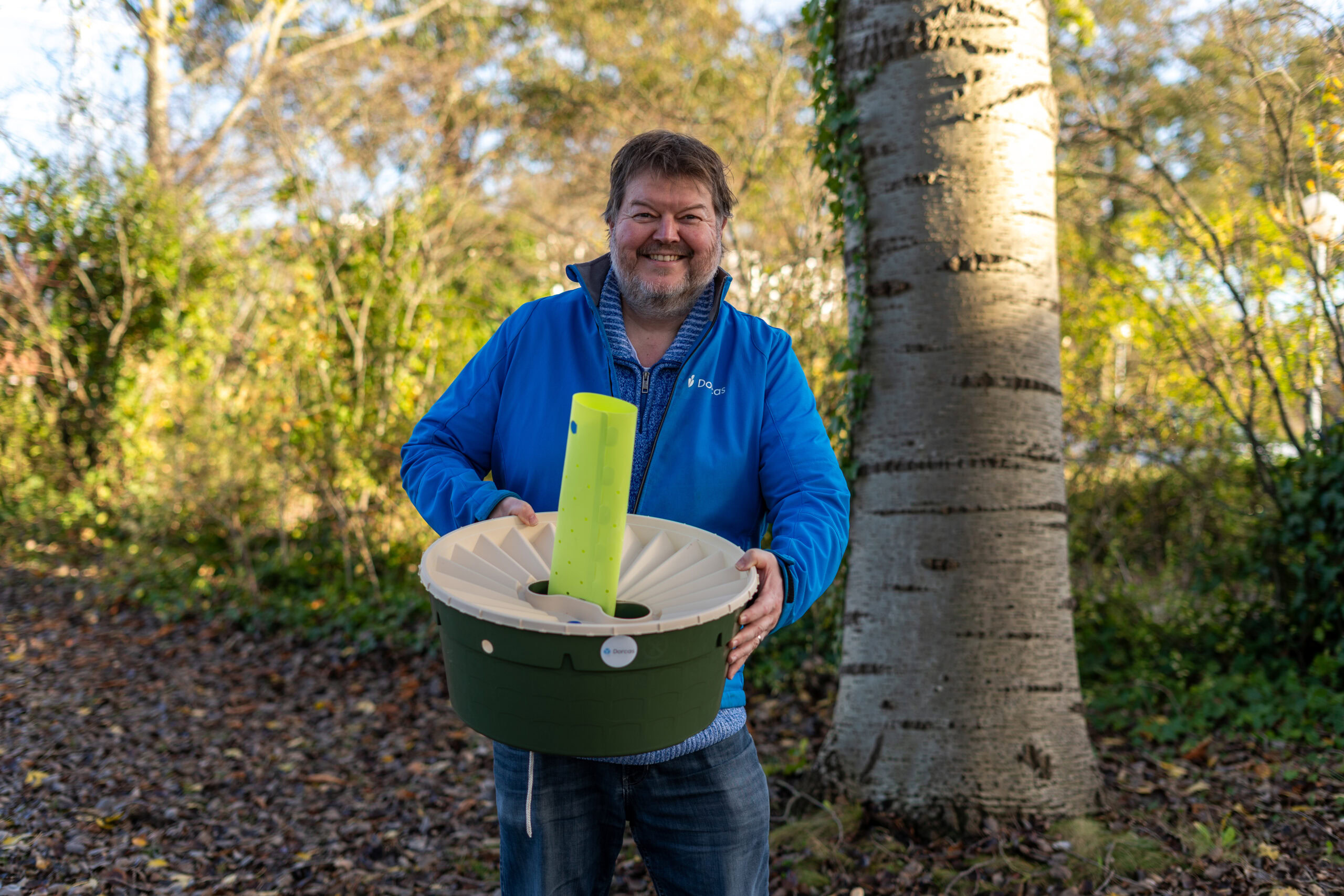 Man met een grote glimlach houdt een groene en witte container met een grote groene pen erin, buiten in een bosrijke omgeving met bomen en herfstkleuren.
