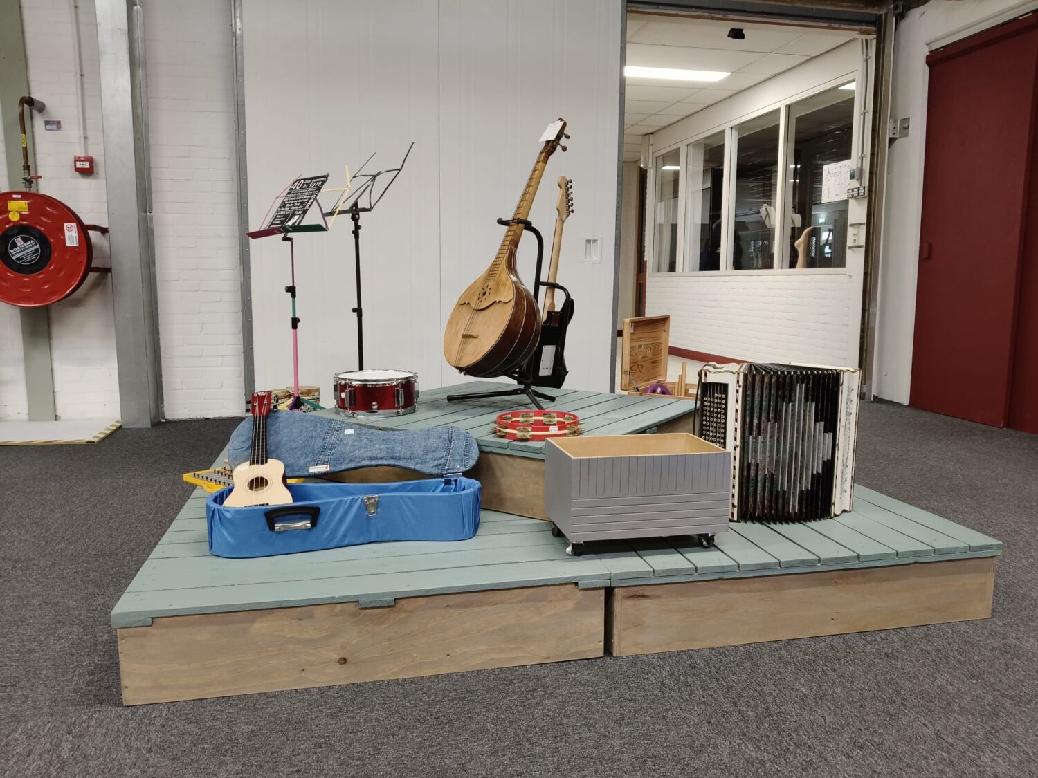 Een verzameling muziekinstrumenten, waaronder gitaren, een drumstel, en bladmuziek, gepresenteerd op een verhoogd platform in een binnenruimte, klaar voor een optreden of oefensessie.