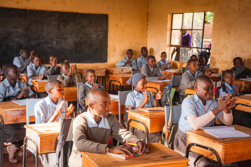 Leerlingen in een klaslokaal met schooluniformen, aandachtig en betrokken bij de les, met een zwarte schoolbord en grote ramen voor natuurlijk licht.