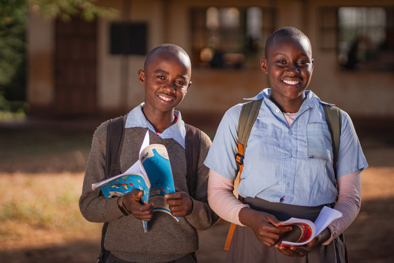 Twee lachende schoolkinderen met boeken en rugzakken buiten op schoolplein.