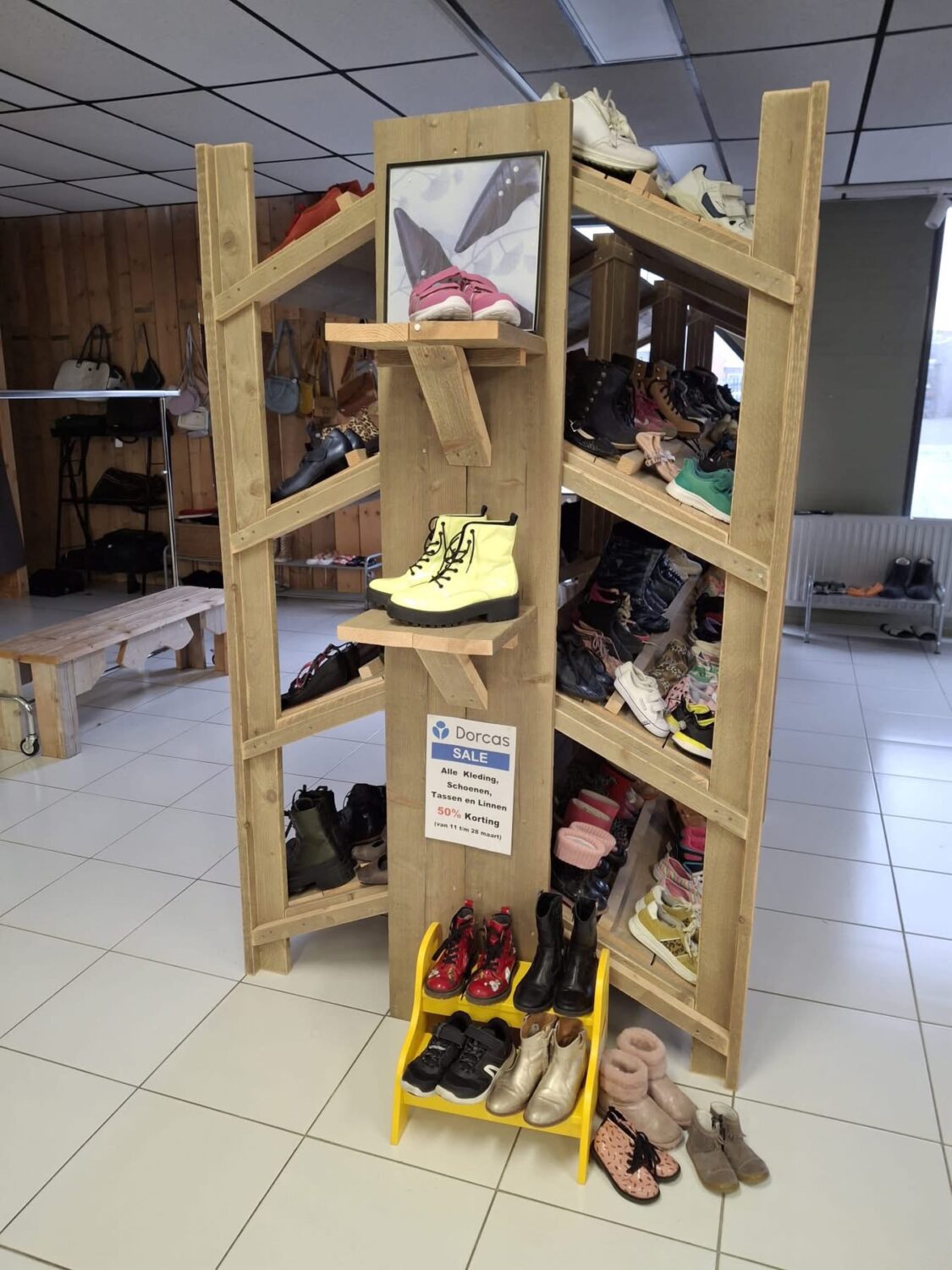 Schoenenwinkel met houten display en diverse schoenen, inclusief kinderschoenen, sneakers en laarzen, in een winkelruimte met witte tegels en houten wand.