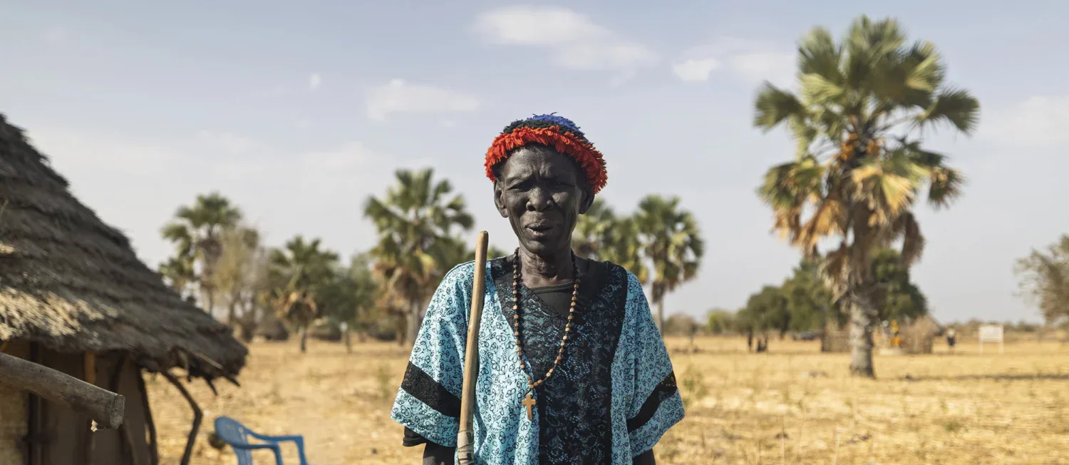 Een oudere vrouw in traditionele kleding staat in een dorre, open omgeving met palmbomen op de achtergrond, onder een heldere blauwe hemel.
