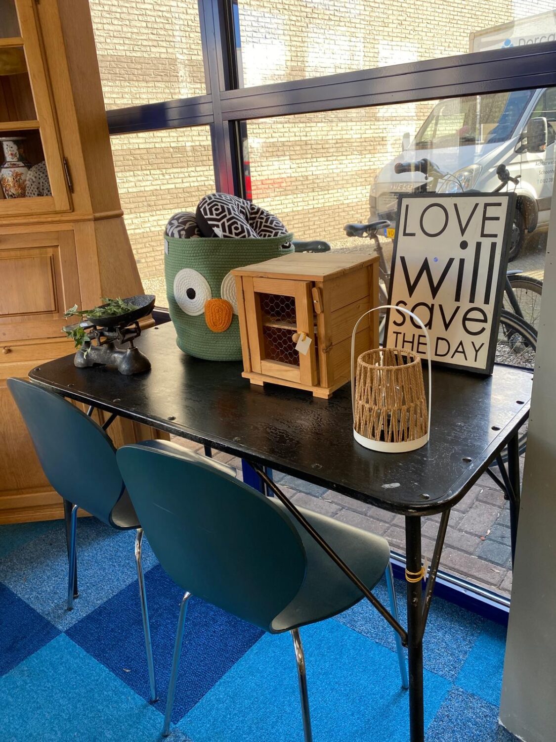 Een tafel met diverse decoratieve items, waaronder een houten kastje, een plantenpot met een owl-ontwerp, een vaas en een canvas met de tekst "Love will save the day" in een gezellige, natuurlijke set.