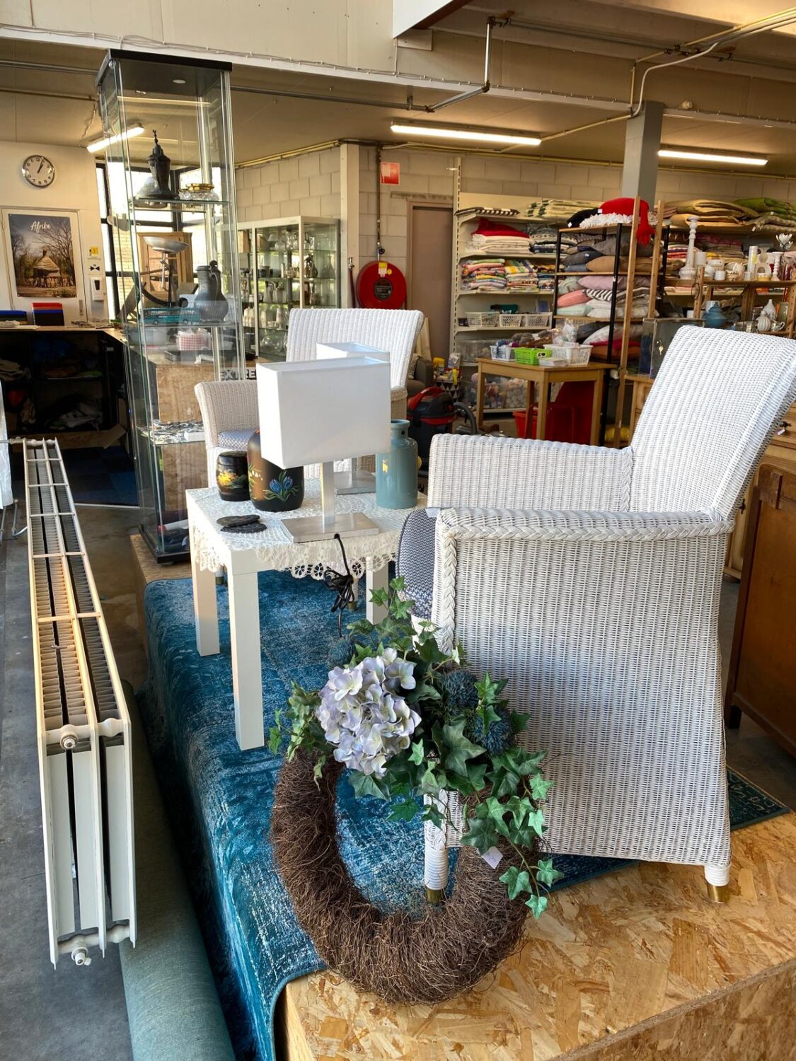 Een gezellige vintage interieur setting met een witte fauteuil, bloemen, kaarsen en decoratieve items, gelegen in een winkel of opslagruimte met diverse stoffen en meubels op de achtergrond.