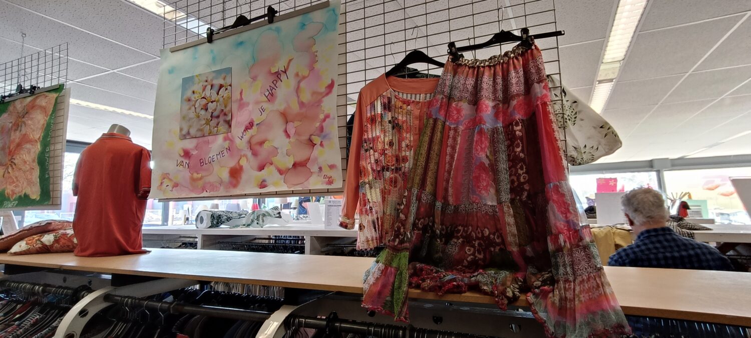 Handgemaakte textielproducten en kleurrijke stoffen hangen in een winkel, met mensen op de achtergrond, passend bij de missie van Dorcas om lokale ambachten te ondersteunen.