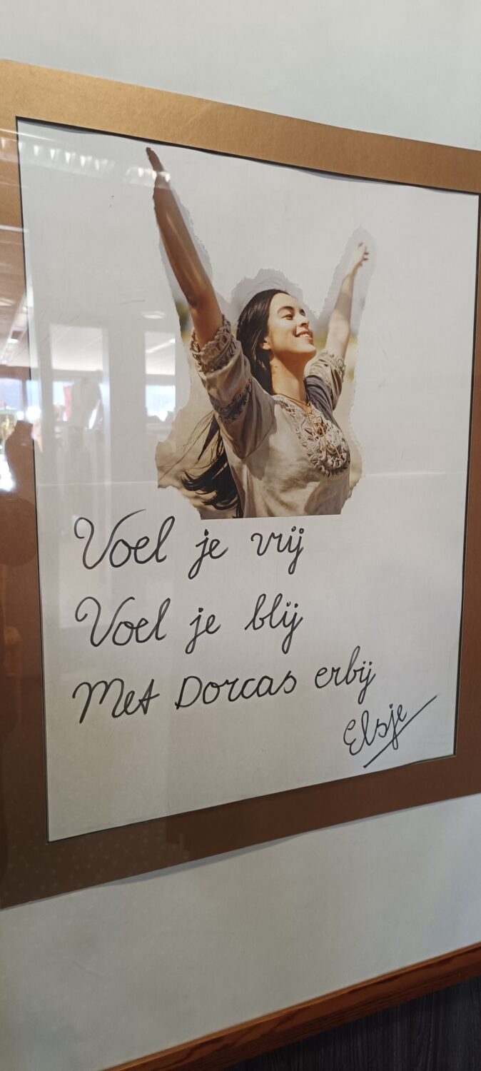 Een vrouw met haar armen omhoog en een glimlach, met een inspirerende boodschap op een whiteboard die zegt: "Voel je vrij, Voel je blij, Met Dorcas erbij".