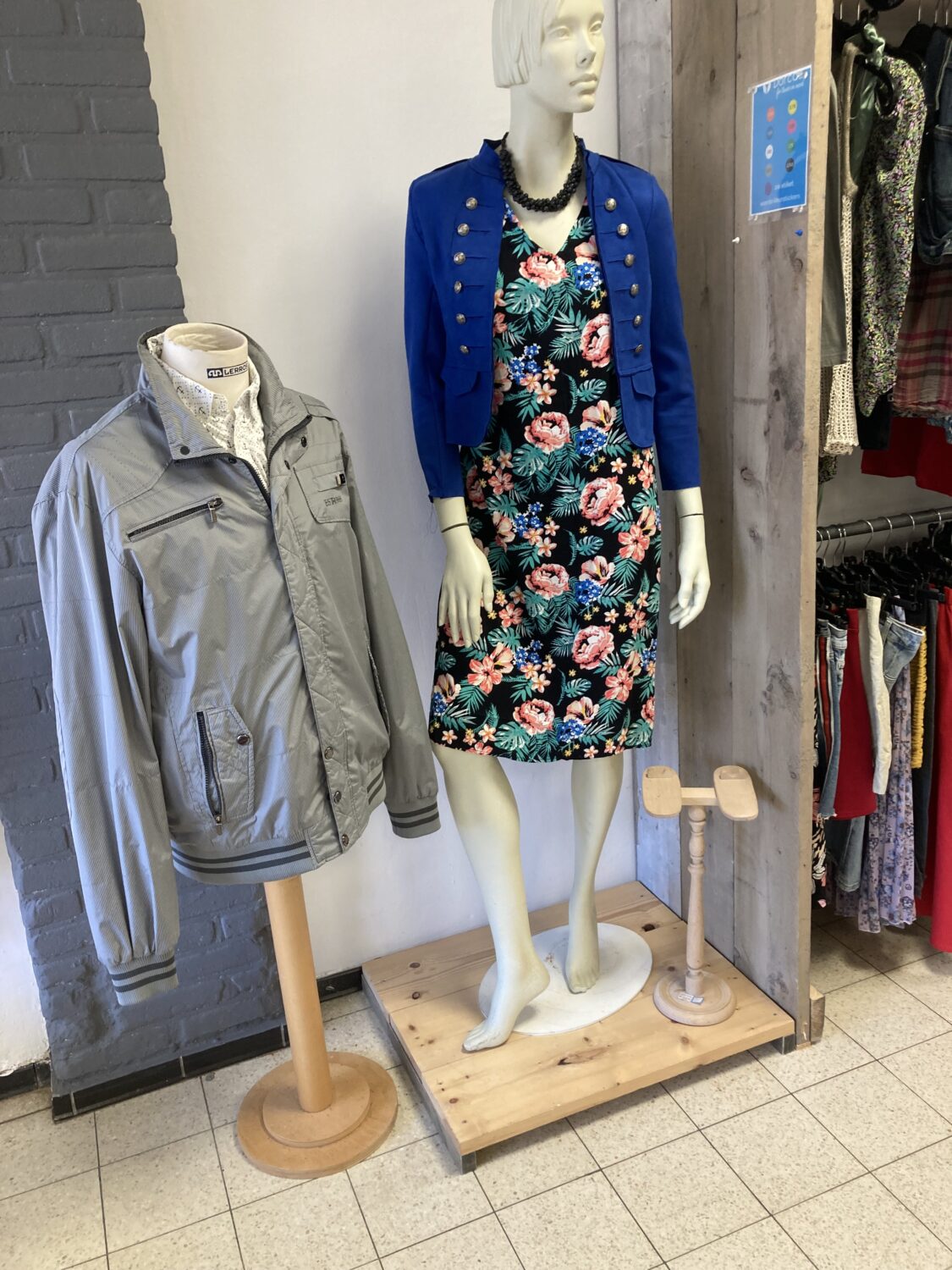 Mannequin met bloemenjurk en blauwe cardigan, tentoongesteld in een winkel, met een grijze jas op een paspop naast haar, geschikt voor modieuze en duurzame kledingkeuzes.