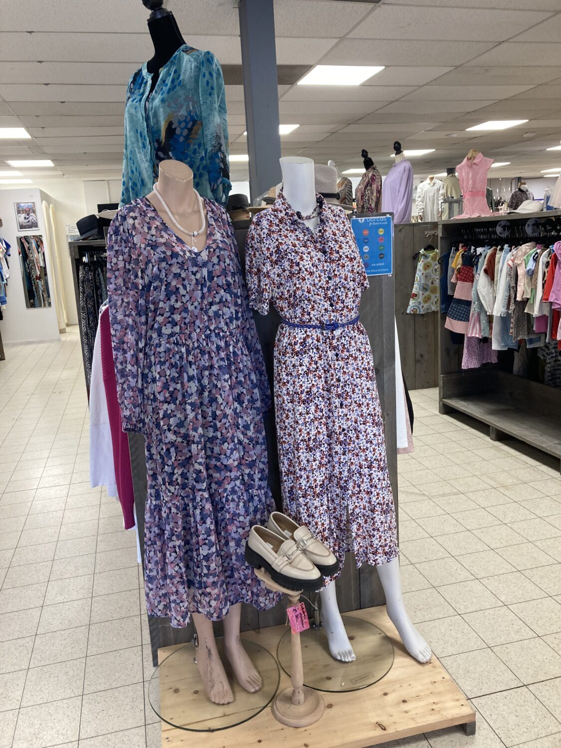Een display met twee vintage bloemenjurken op mannequins, omringd door andere kledingstukken in een winkel.