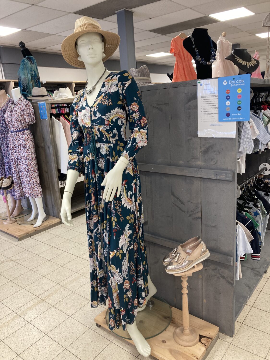 Een mannequin draagt een lange, bloemenprint zomerjurk met een strohoed in een winkel van Dorcas, omringd door andere kledingstukken en accessoires.